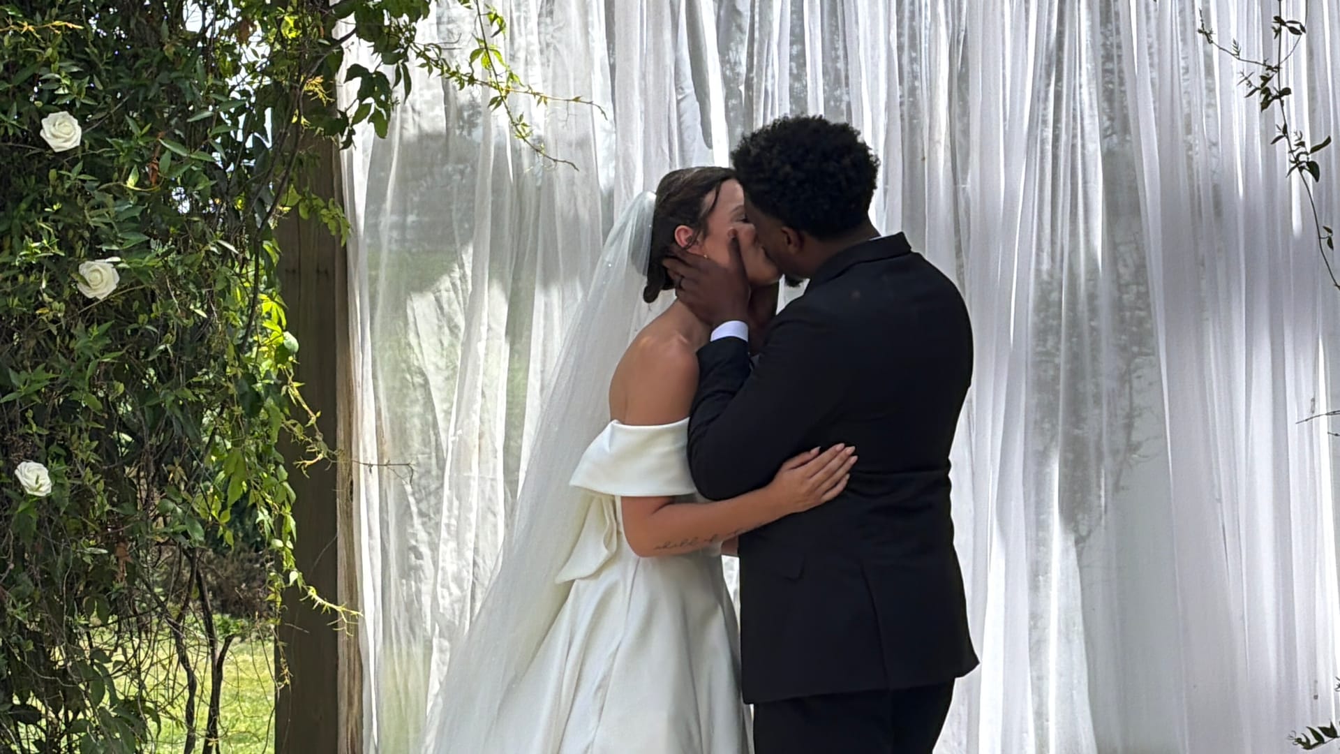 Bride in off-shoulder white gown and veil shares first kiss with groom in black suit beneath flowing white draping and greenery floral arch at Scarborough House wedding ceremony.
