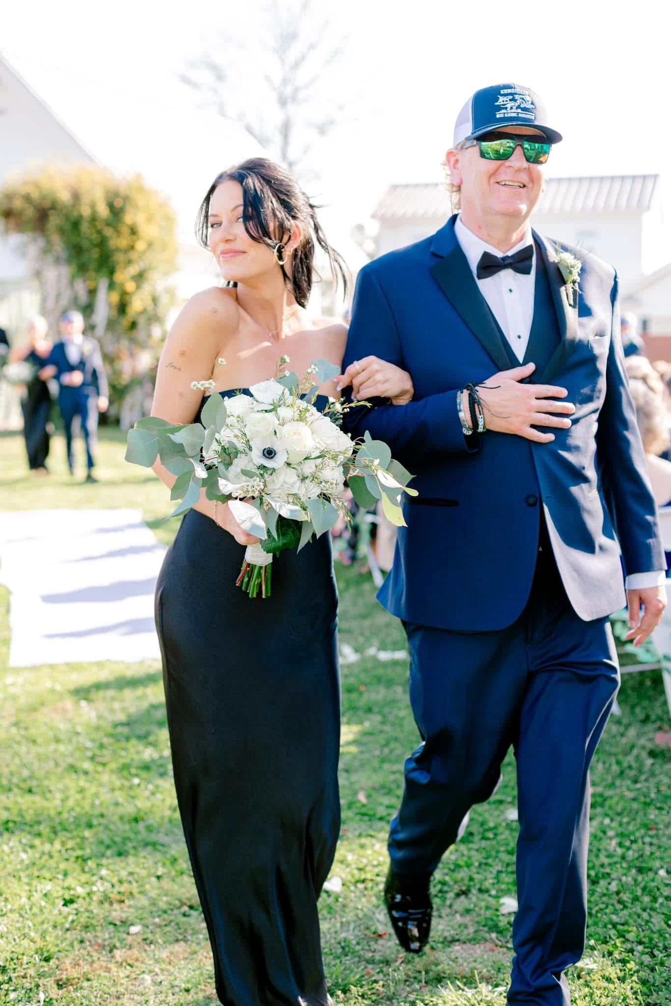Bride in a strapless black gown holding a white floral bouquet walks down the outdoor ceremony aisle arm-in-arm with her escort in a navy tuxedo at Scarborough House.
