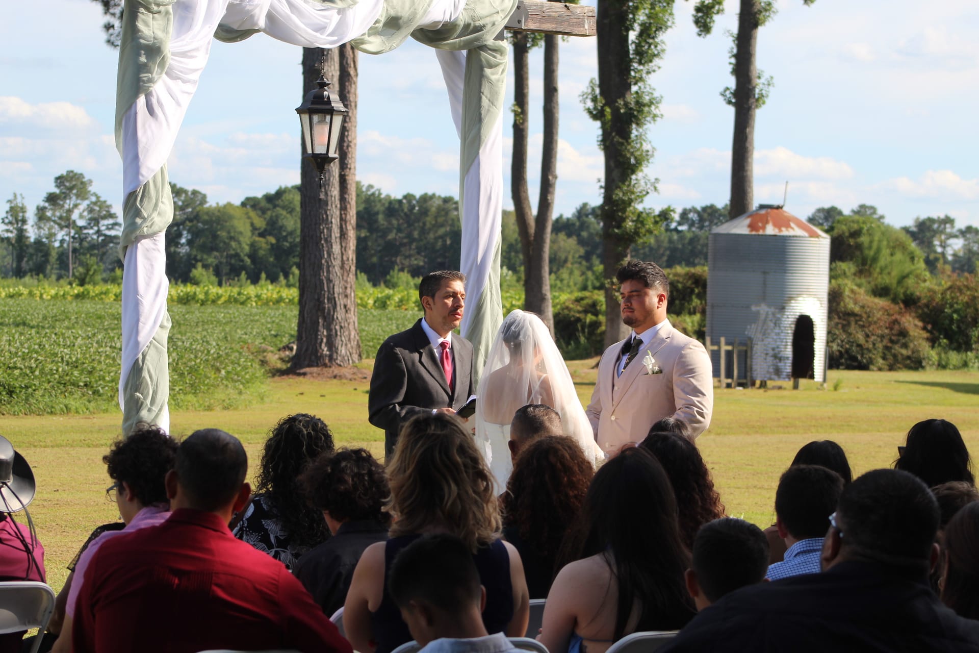 Outdoor wedding ceremony on the estate lawn as a bride in a white veil and groom in a light suit exchange vows beneath a draped wooden arch with farmland fields and trees in the background.
