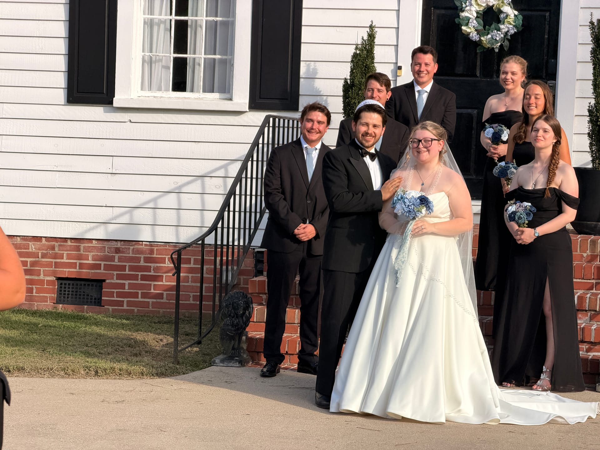 Bride in a white ballgown and groom in a black tuxedo pose with their wedding party on the front steps of the historic 1821 Scarborough House manor, bridesmaids in black dresses holding blue bouquets.