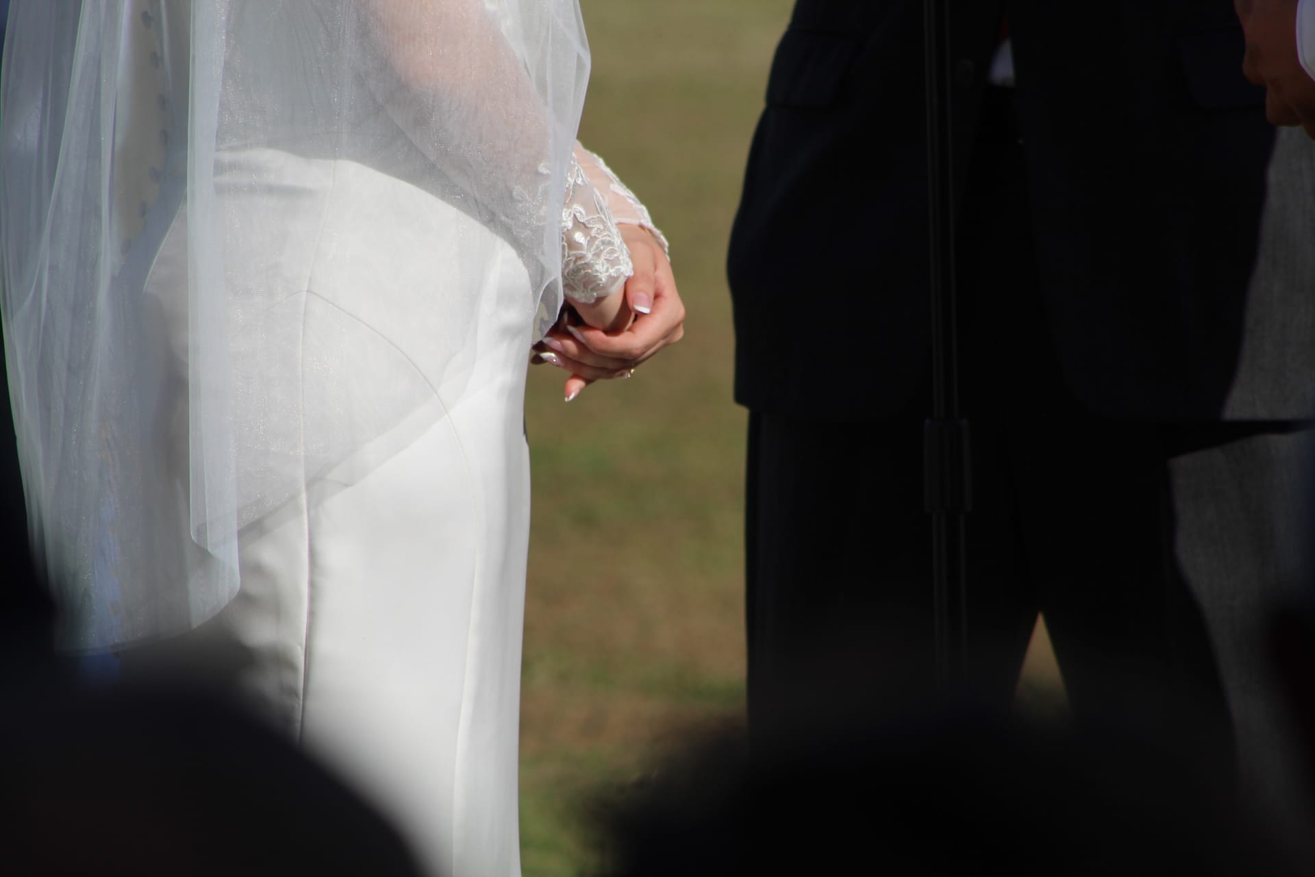 Close-up of a bride in a white lace-sleeved wedding gown gently clasping her hands during an outdoor ceremony on the estate lawn.