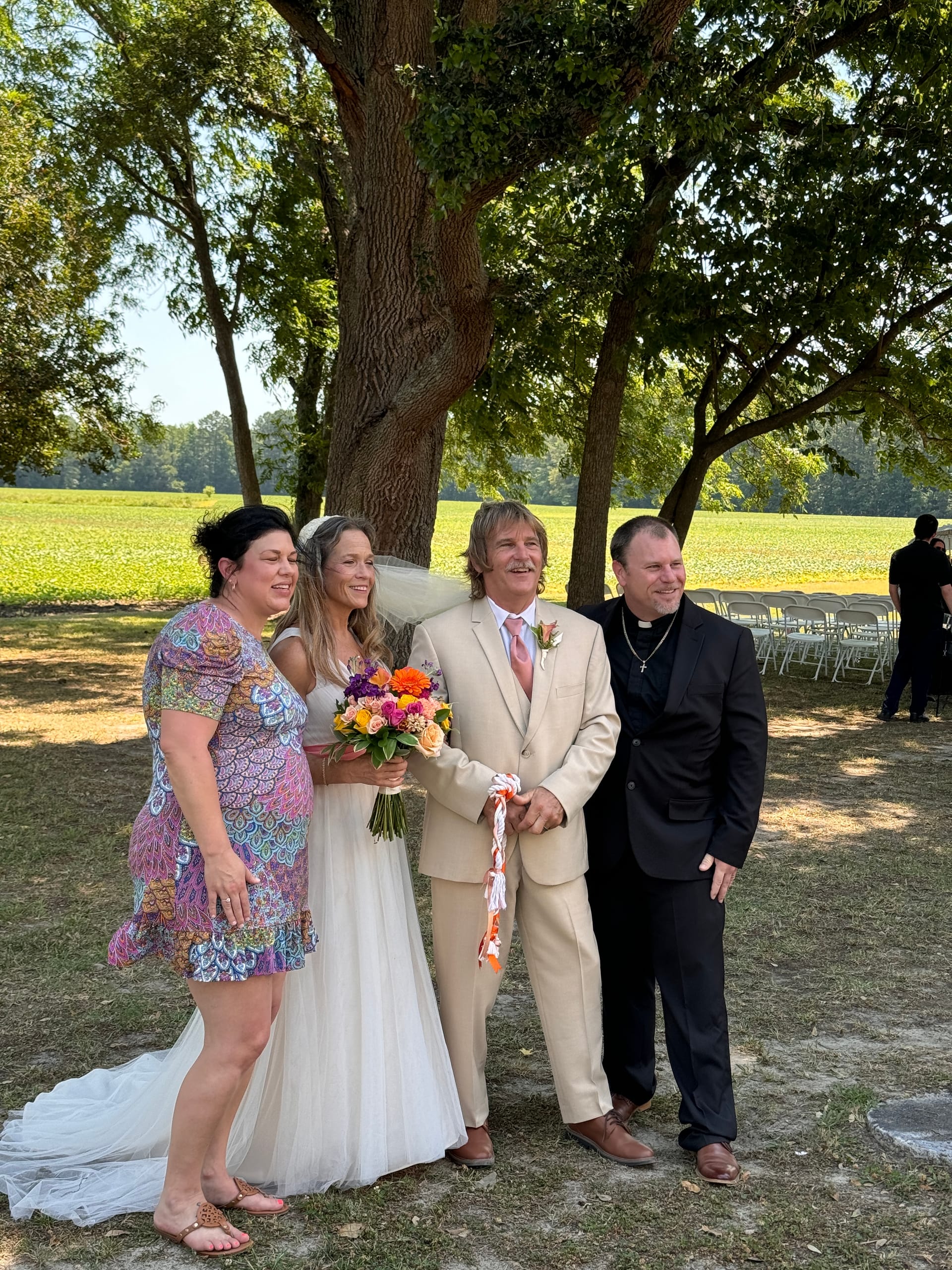 Bride and groom smile with two guests including an officiant beneath large oak trees on the ceremony lawn at Scarborough House estate, with open farmland fields visible in the background.