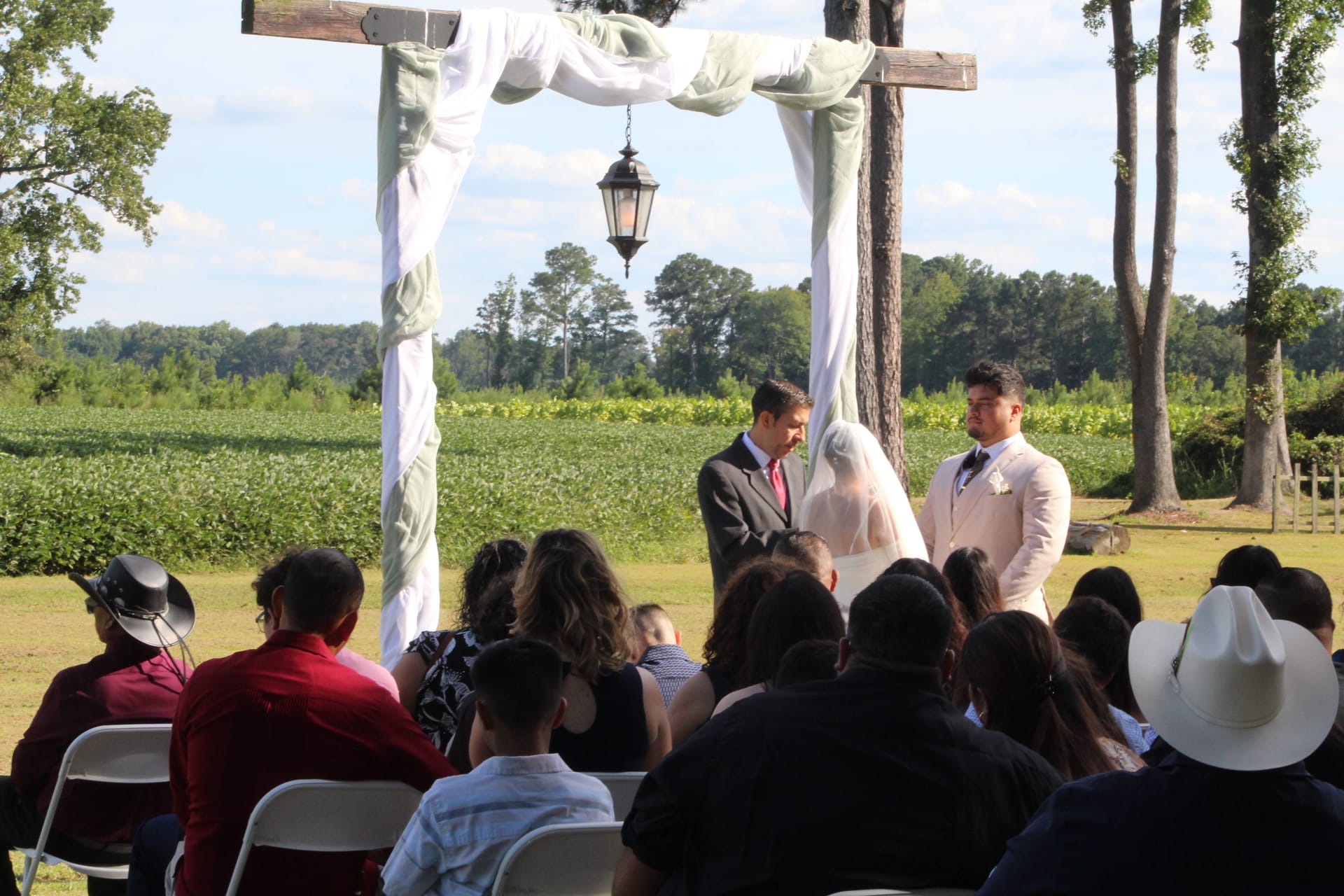 Wedding ceremony on the outdoor lawn at Scarborough House with the couple exchanging vows beneath a rustic wooden arch draped in white fabric and a hanging lantern, surrounded by seated guests and a backdrop of green farmland fields and tall trees.