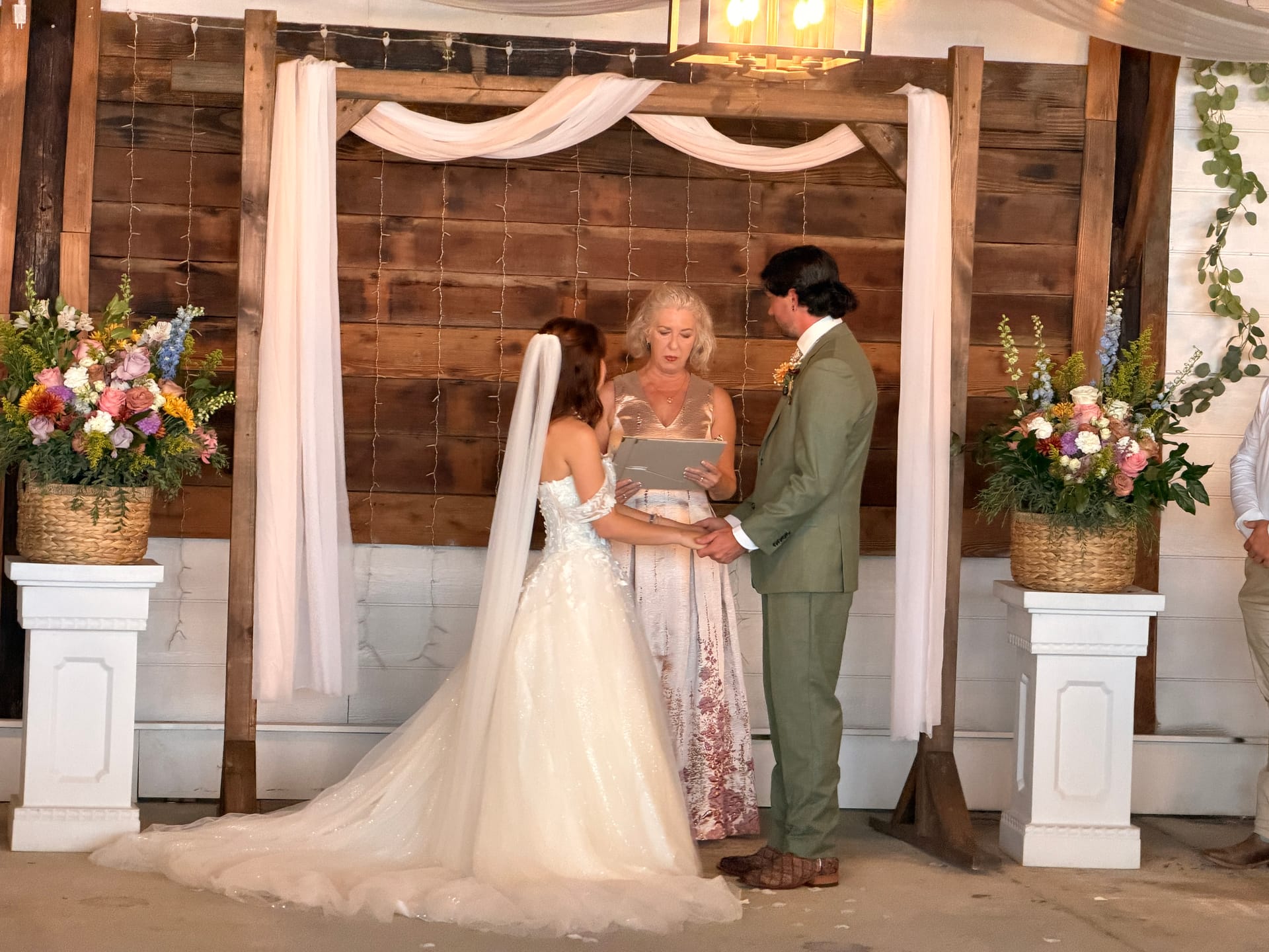 Bride in a flowing white gown and groom in a sage suit exchange vows beneath a rustic wooden arch adorned with white draping and colorful floral arrangements inside the Scarborough House venue.