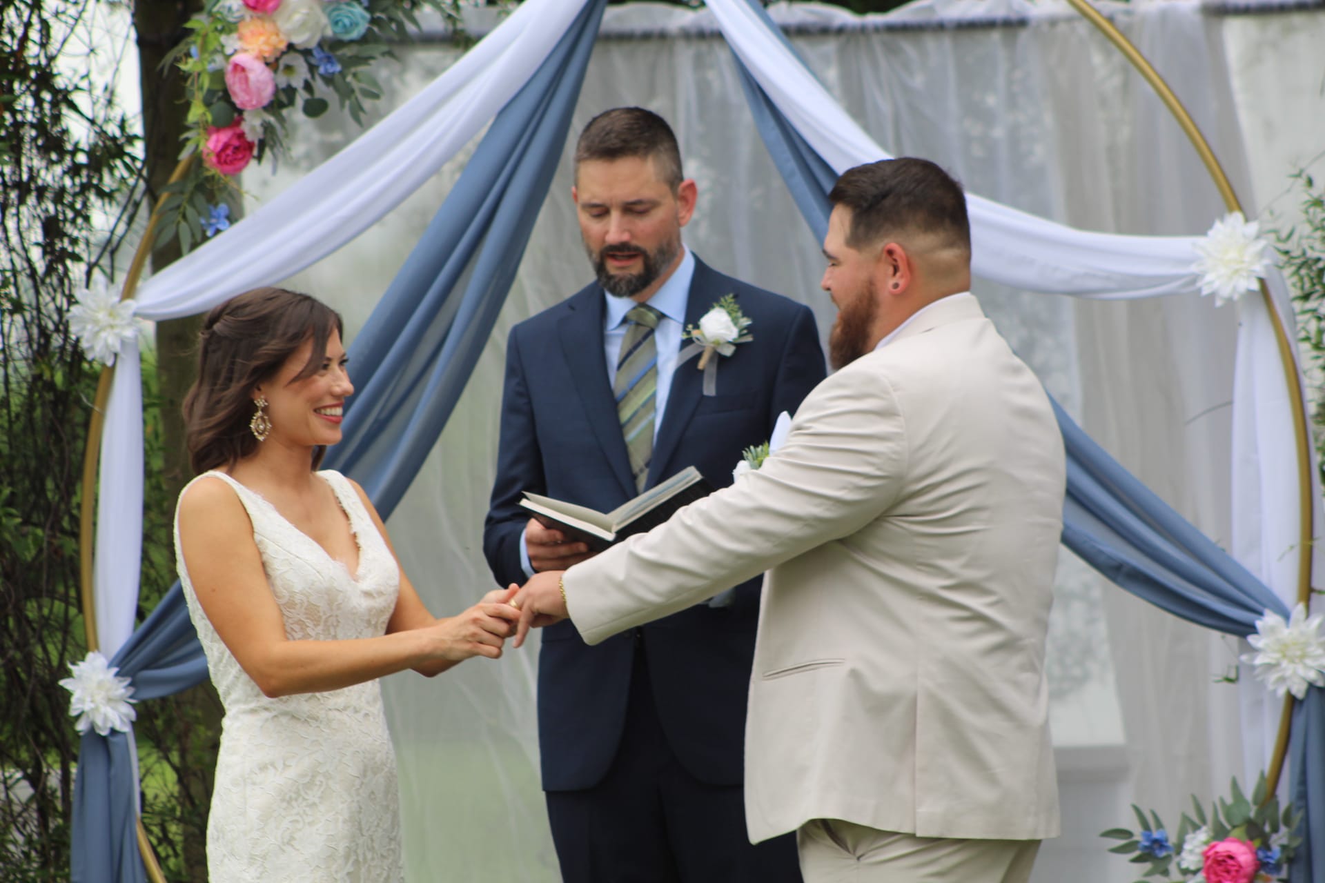 Smiling bride in a lace gown holds hands with the groom in a tan suit during their outdoor ring exchange ceremony beneath a floral-draped circular arch at Scarborough House.