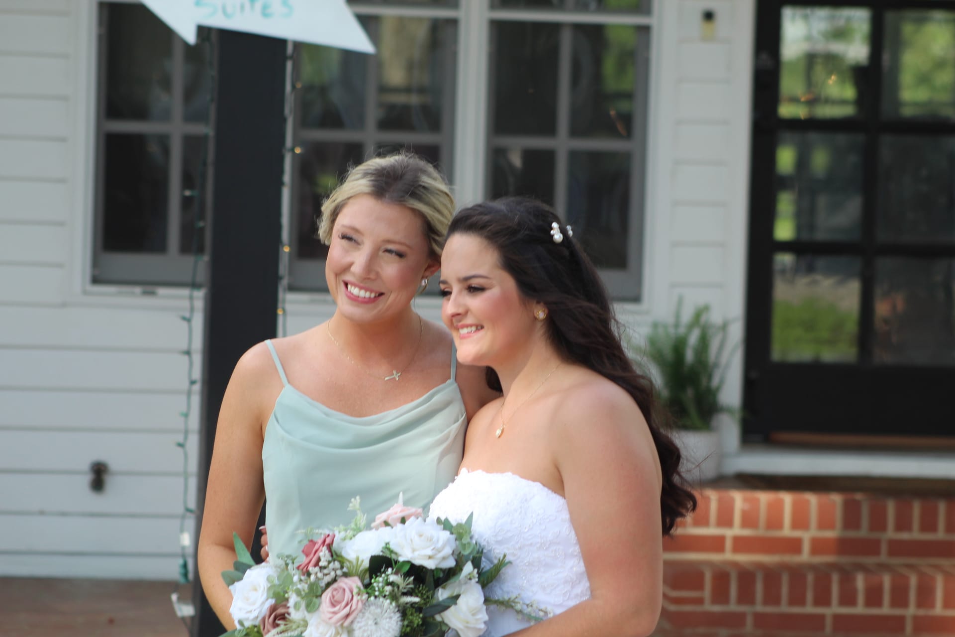 Smiling bride in white strapless gown holding pink and white bouquet poses with bridesmaid in sage green dress in front of the historic Scarborough House manor