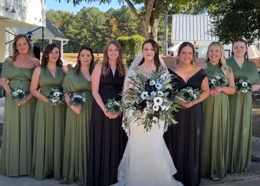 Bride in a white gown holding a large black and white floral bouquet stands with seven bridesmaids in sage green and black dresses outdoors under oak trees at Scarborough House wedding venue.