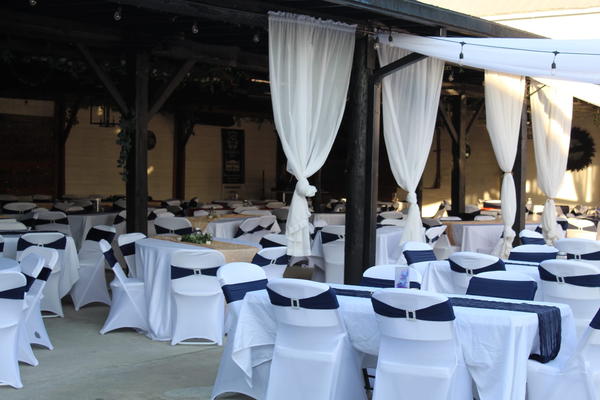 Elegantly decorated outdoor reception pavilion at Scarborough House featuring white chair covers with navy sashes, draped white chiffon curtains on dark wooden columns, and round tables with burlap and greenery centerpieces.