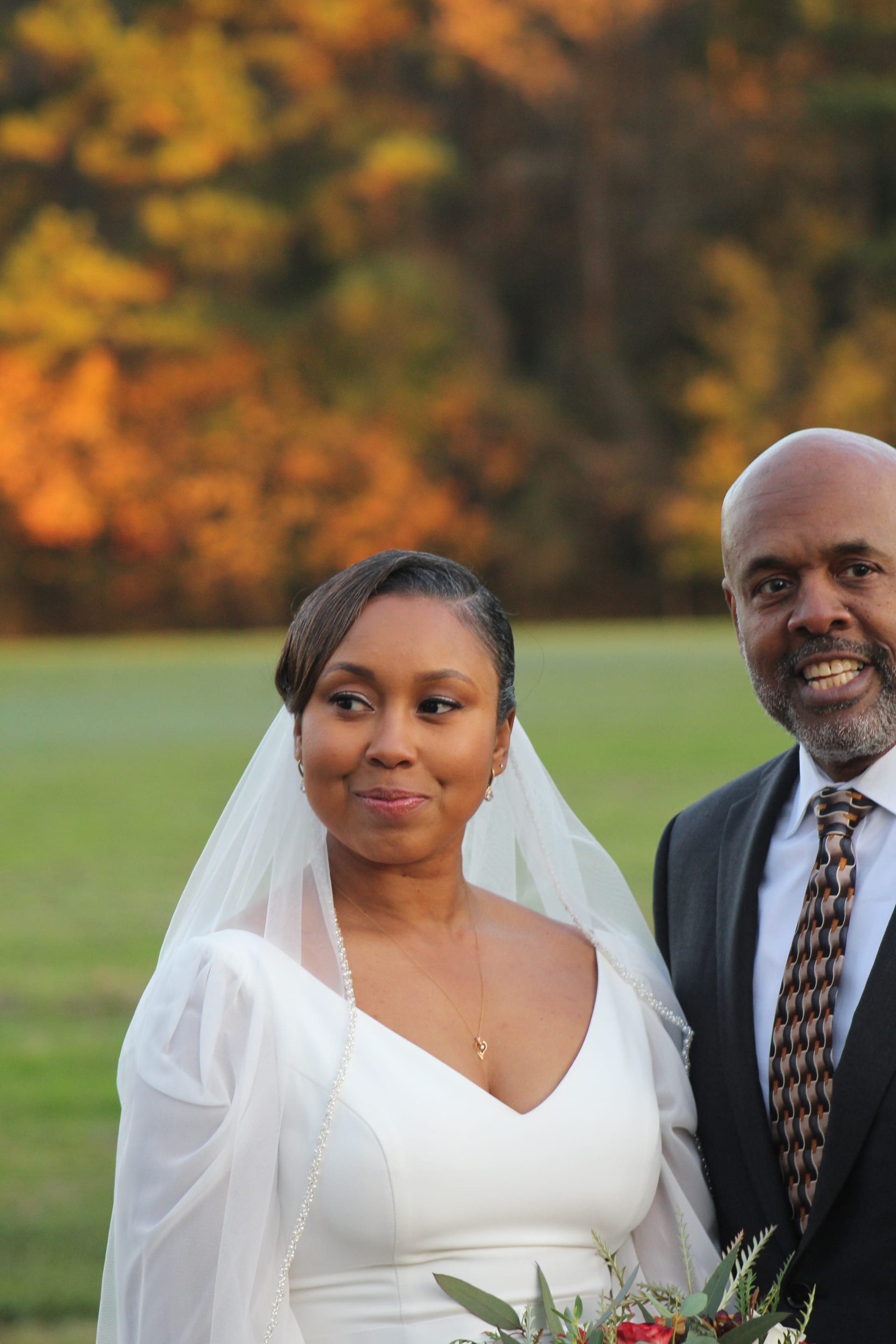A smiling bride in a white gown and veil stands beside a gentleman in a dark suit and tie against a backdrop of vibrant autumn foliage on the ceremony lawn at Scarborough House.