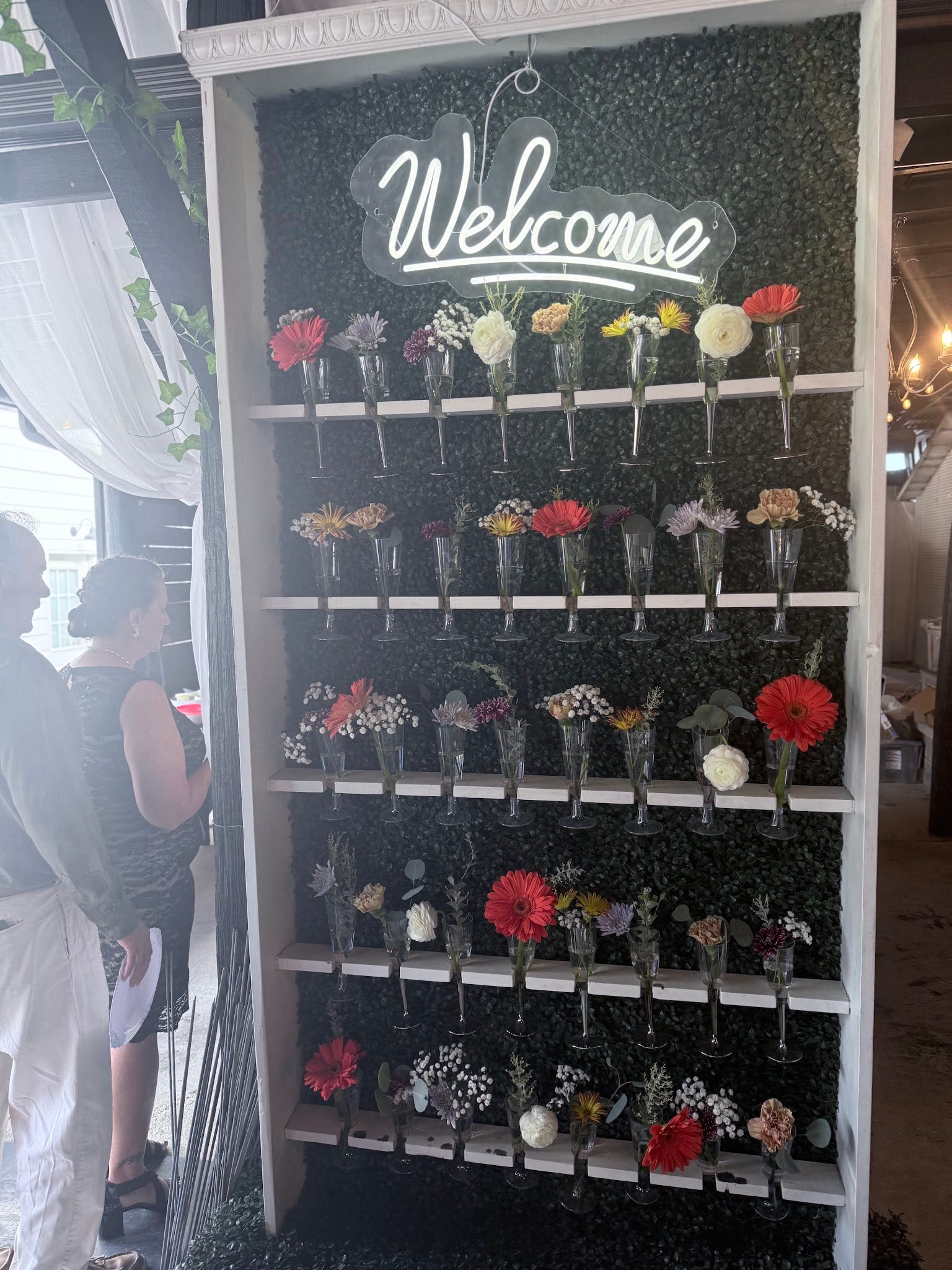A glowing neon Welcome sign anchors a lush boxwood wall lined with white shelves holding champagne flutes filled with colorful fresh flowers at a wedding reception.