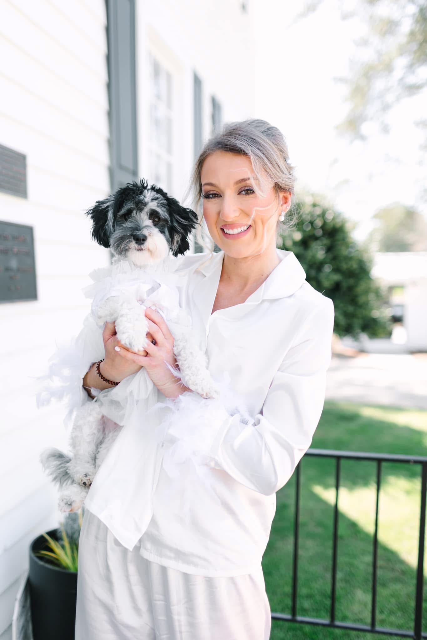 Smiling bride in a white shirt holding a small black and white dog dressed in a white tulle outfit outside the historic Scarborough House manor.