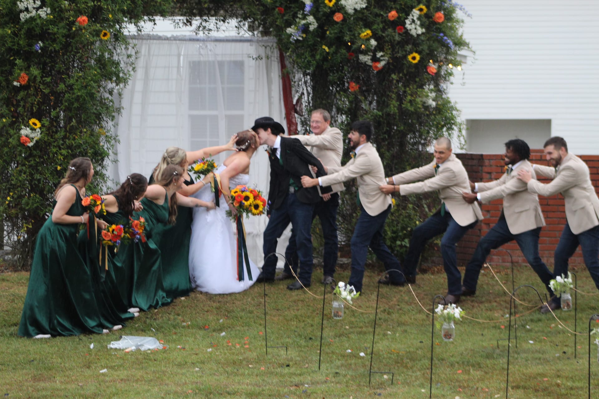 Bridesmaids in emerald green gowns and groomsmen in tan blazers playfully tug the bride and groom apart in a tug-of-war pose on the ceremony lawn at Scarborough House, surrounded by sunflower bouquets and a floral arch.