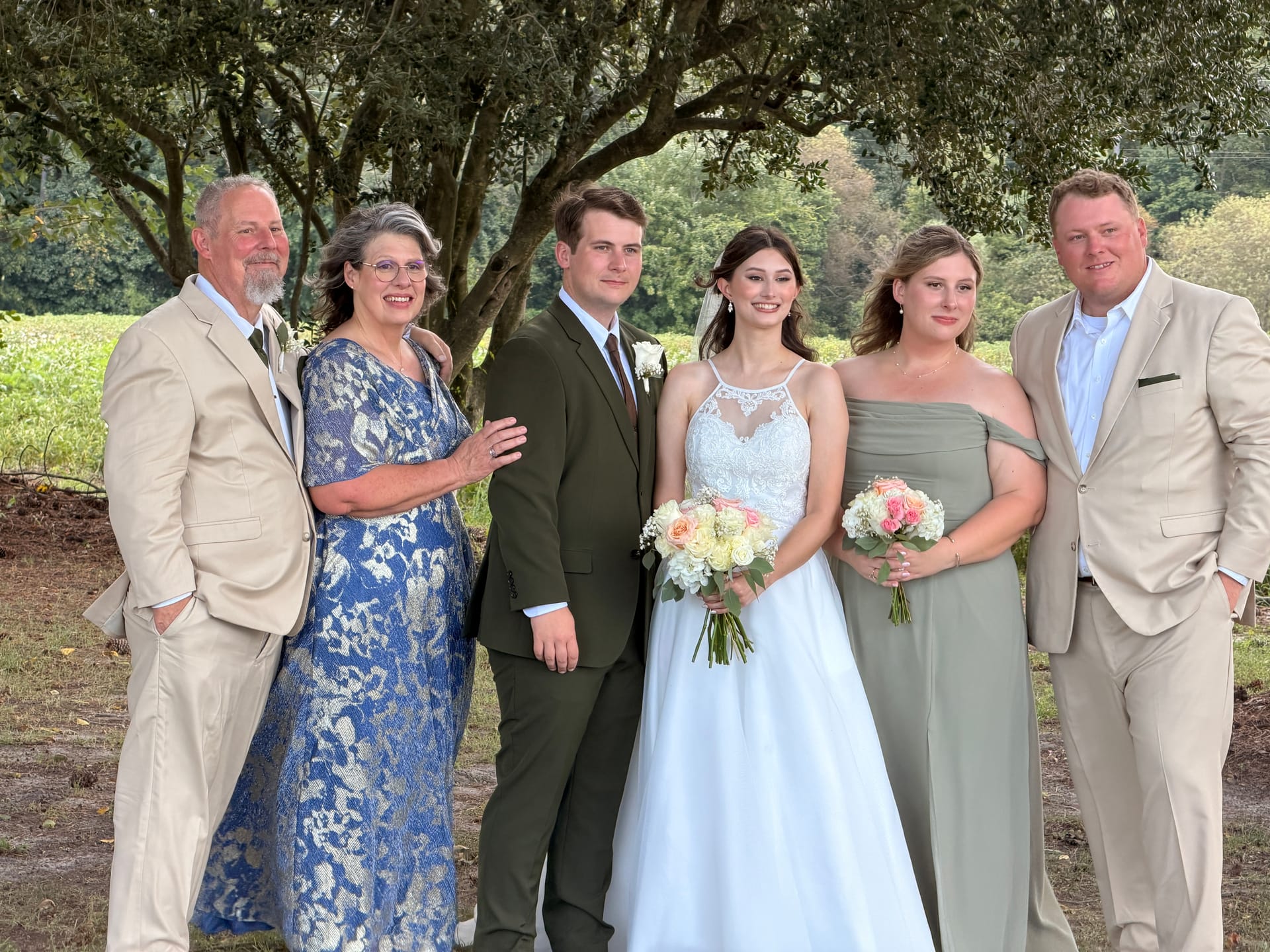 A bride and groom pose with four family members in formal attire beneath oak trees with lush farmland fields in the background at Scarborough House.