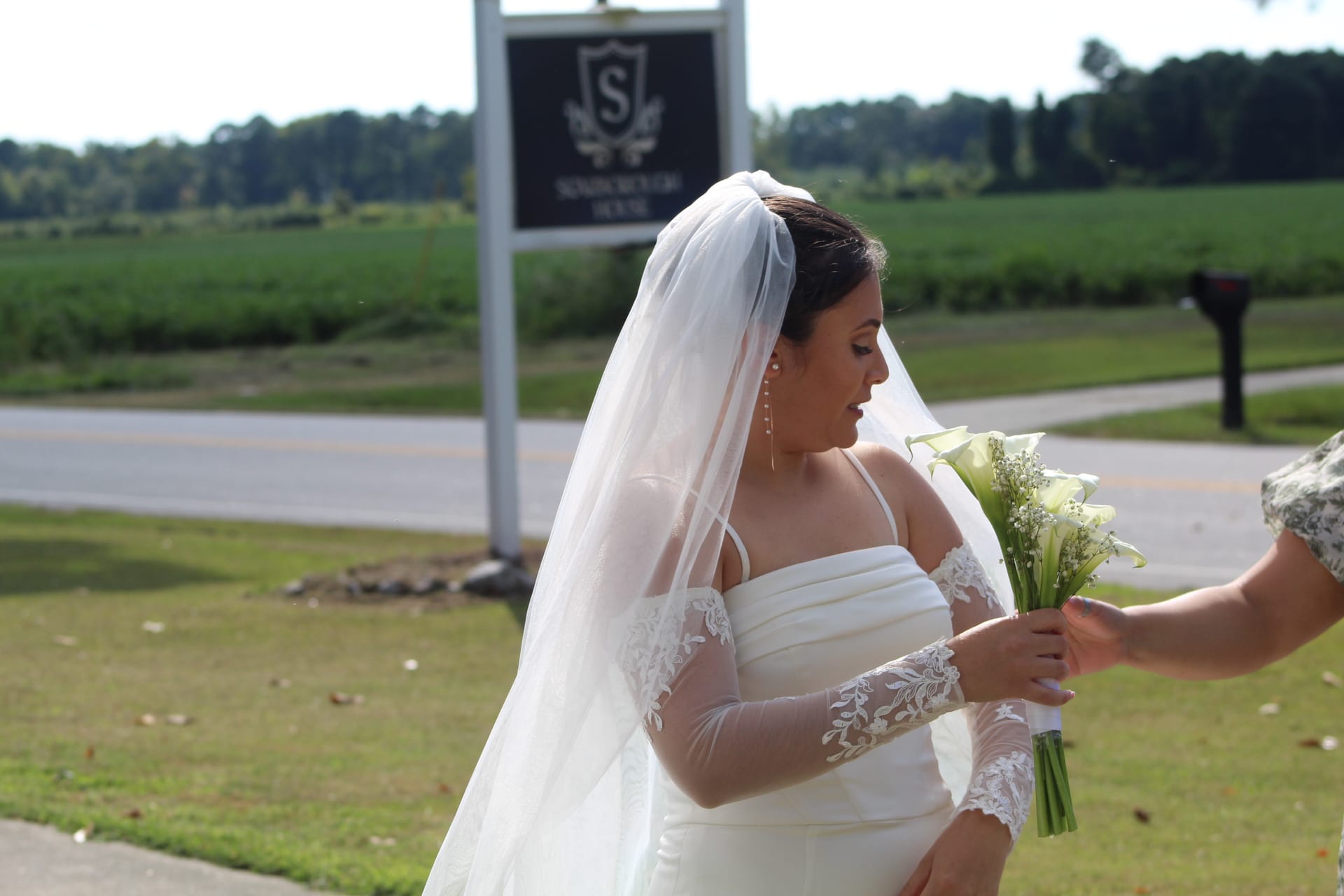 Bride in a white lace-sleeve gown and veil receives her white calla lily bouquet near the Scarborough House estate sign on a sunny wedding day.