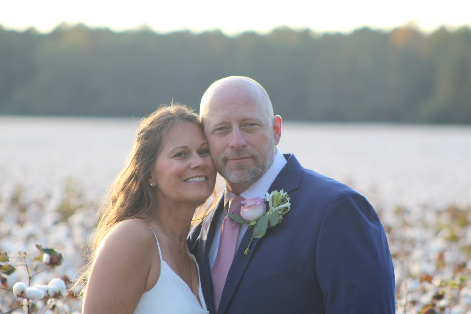 Bride in white gown leans cheek-to-cheek with groom in navy suit and pink boutonniere as golden sunset light glows over a cotton field at Scarborough House.