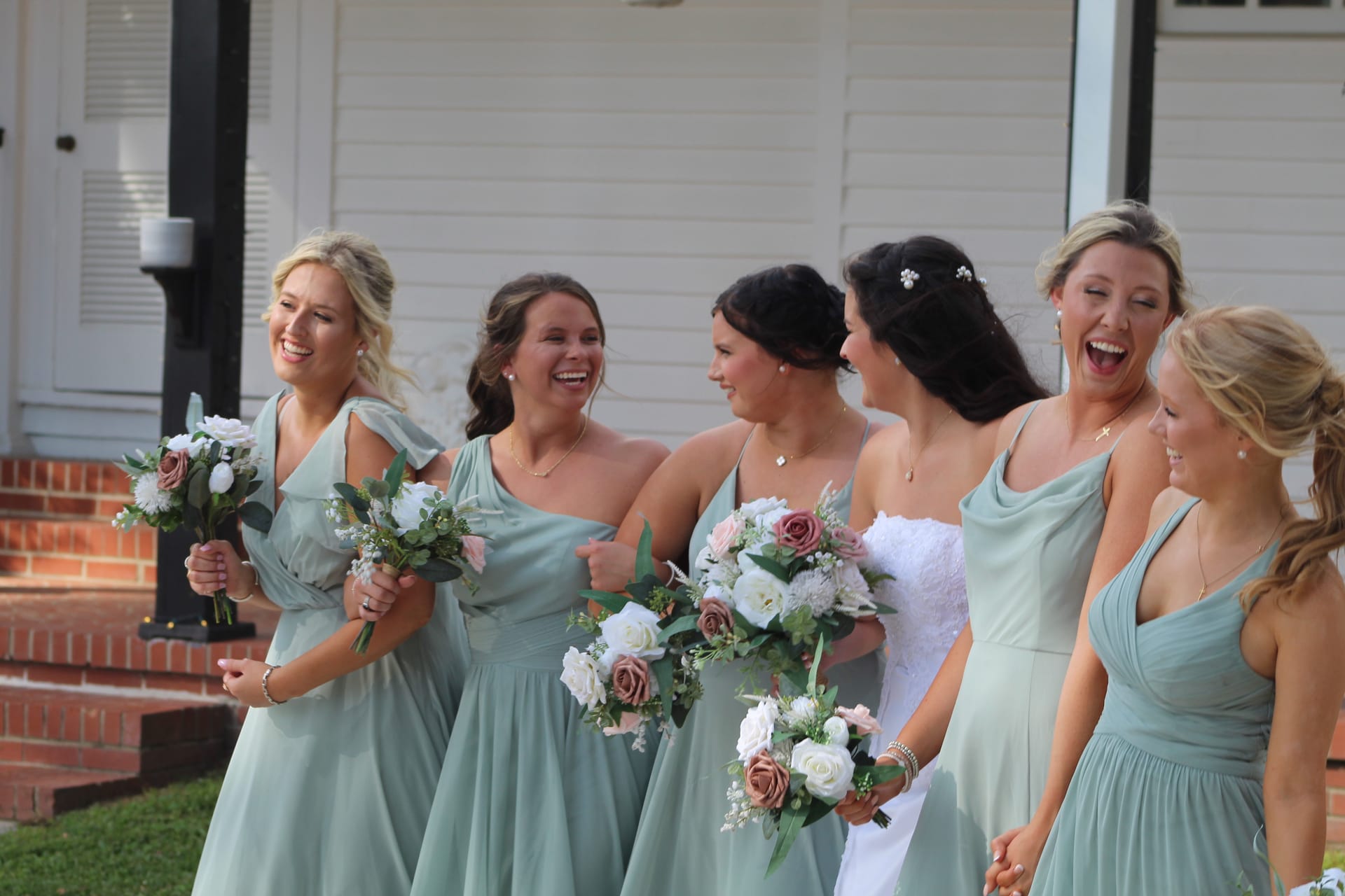 Bride in white gown laughing with five bridesmaids in sage green dresses holding floral bouquets outside a historic white manor house.