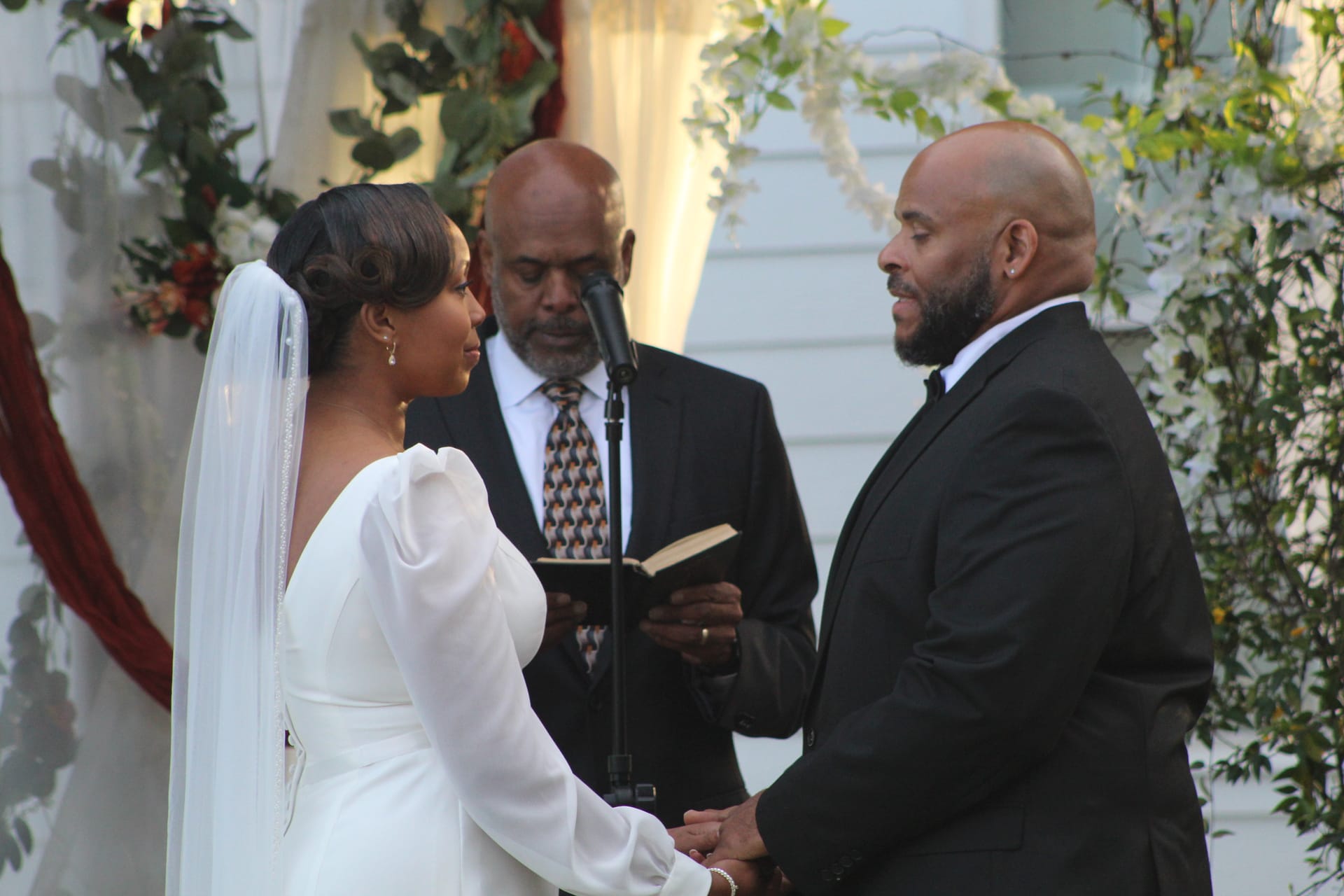 Bride in a long-sleeve white gown and veil holds hands with her groom in a black suit as an officiant reads from a book beneath a floral-draped arch at Scarborough House.
