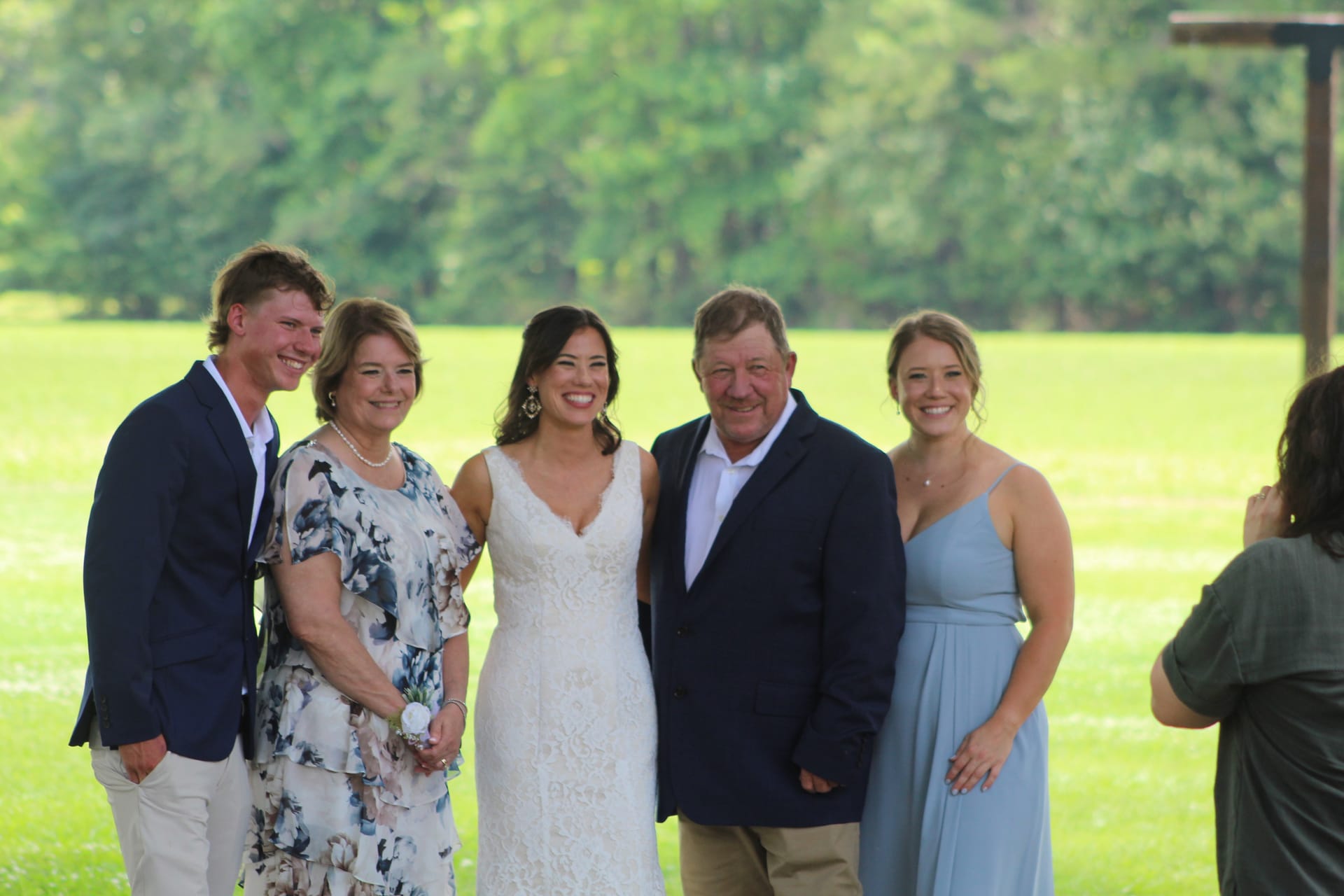 A smiling bride in a white lace gown poses with four family members on the green ceremony lawn at Scarborough House, surrounded by lush trees.