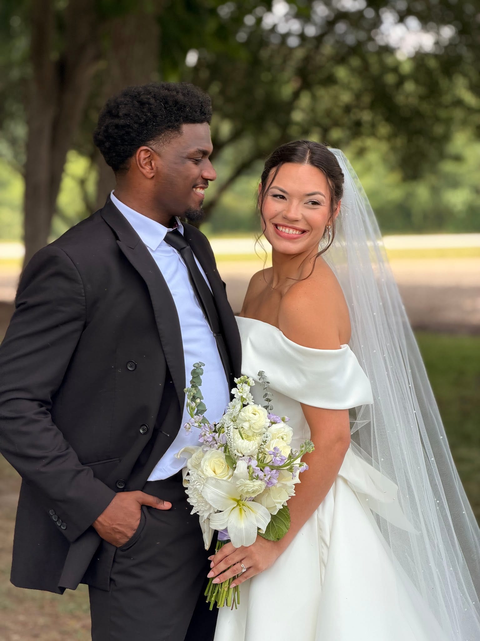 Bride in an off-shoulder white gown holding a white and lavender bouquet smiles brightly beside her suited groom beneath the sprawling oak trees at Scarborough House.