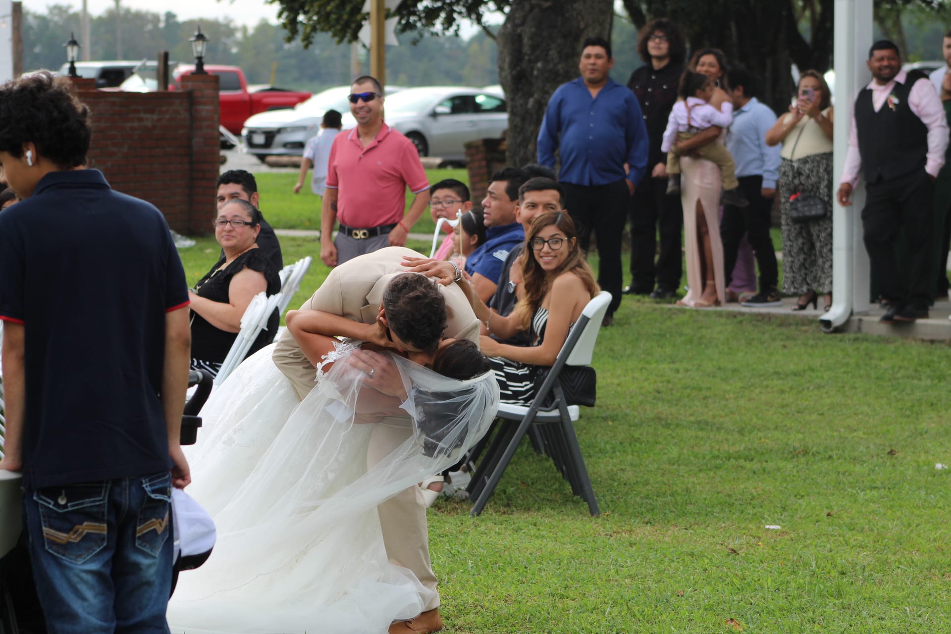 Groom dips bride for a kiss during their outdoor wedding ceremony on the lawn as smiling guests look on.