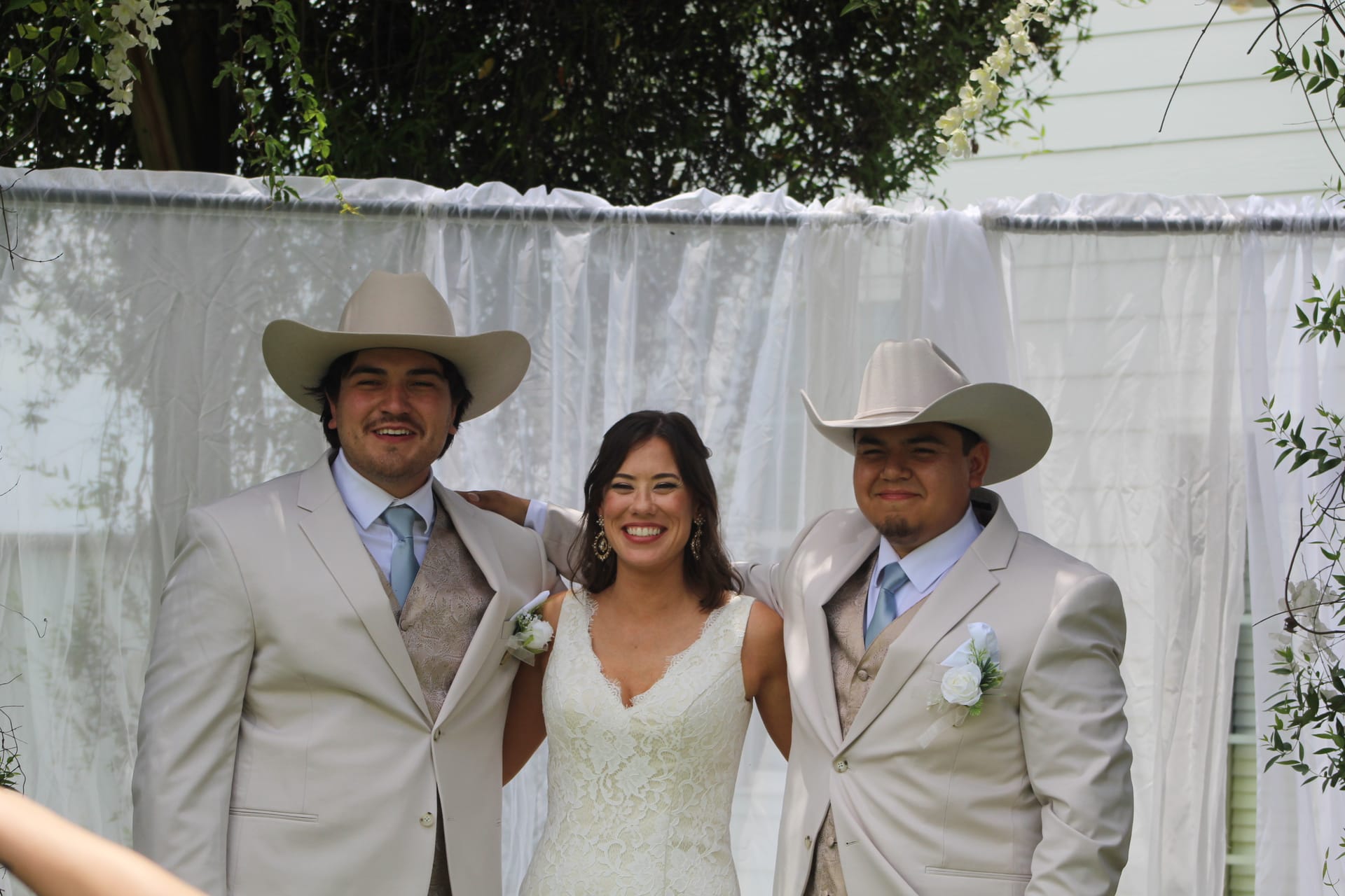 Bride in a lace wedding gown smiles between two groomsmen wearing tan suits and cowboy hats in front of a white draped ceremony backdrop at an outdoor wedding.