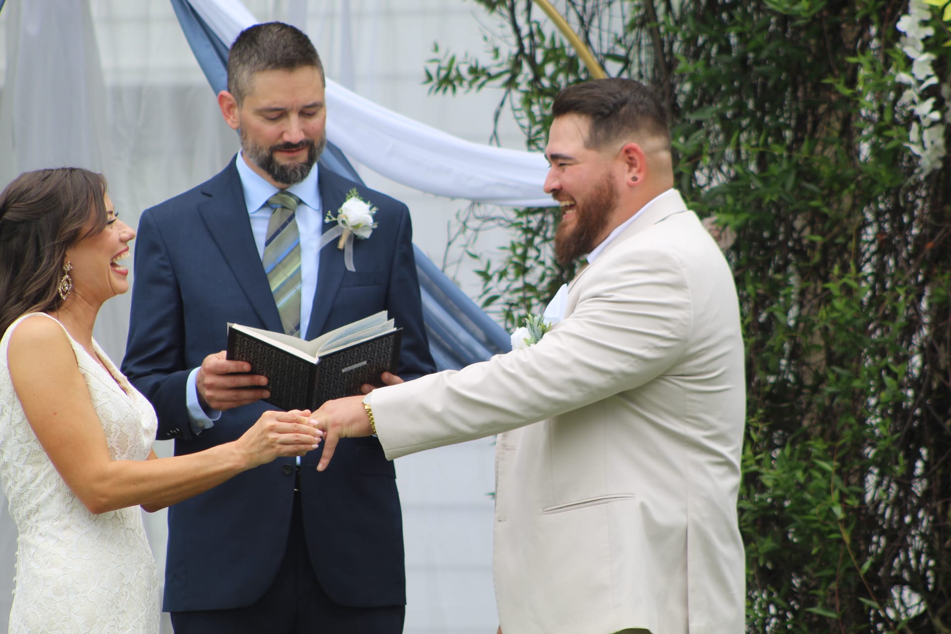 Bride and groom laugh joyfully during their ring exchange ceremony at Scarborough House with a lush greenery arch and draped blue fabric in the background.