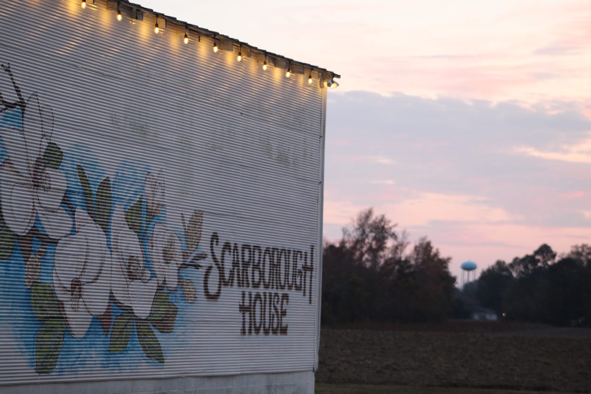 Scarborough House barn exterior adorned with warm string lights glowing against a pink sunset sky with open farmland and trees in the background.