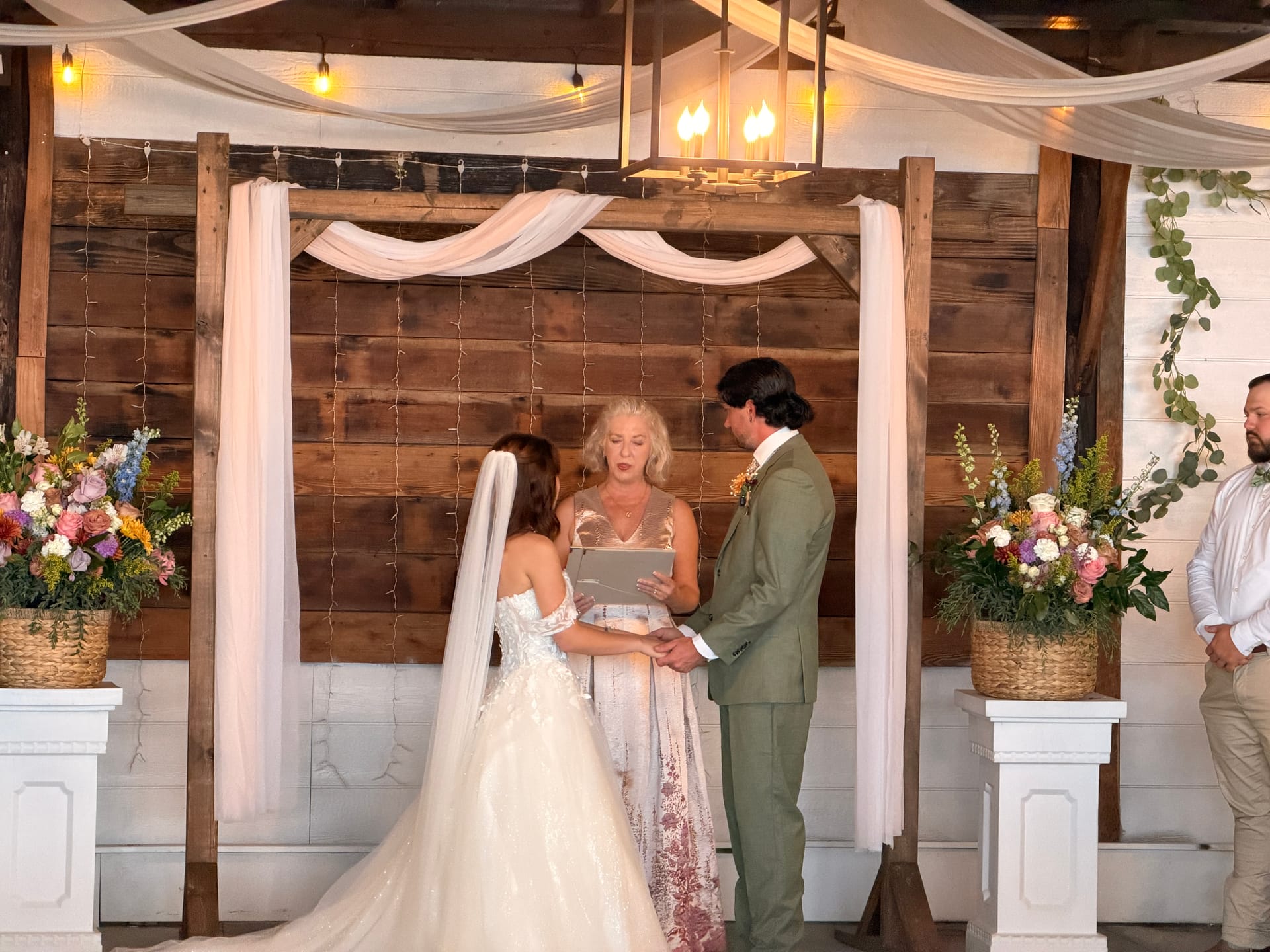 Bride and groom exchange vows before a rustic wood plank altar draped in white fabric and flanked by colorful wildflower arrangements inside the Scarborough House wedding venue.