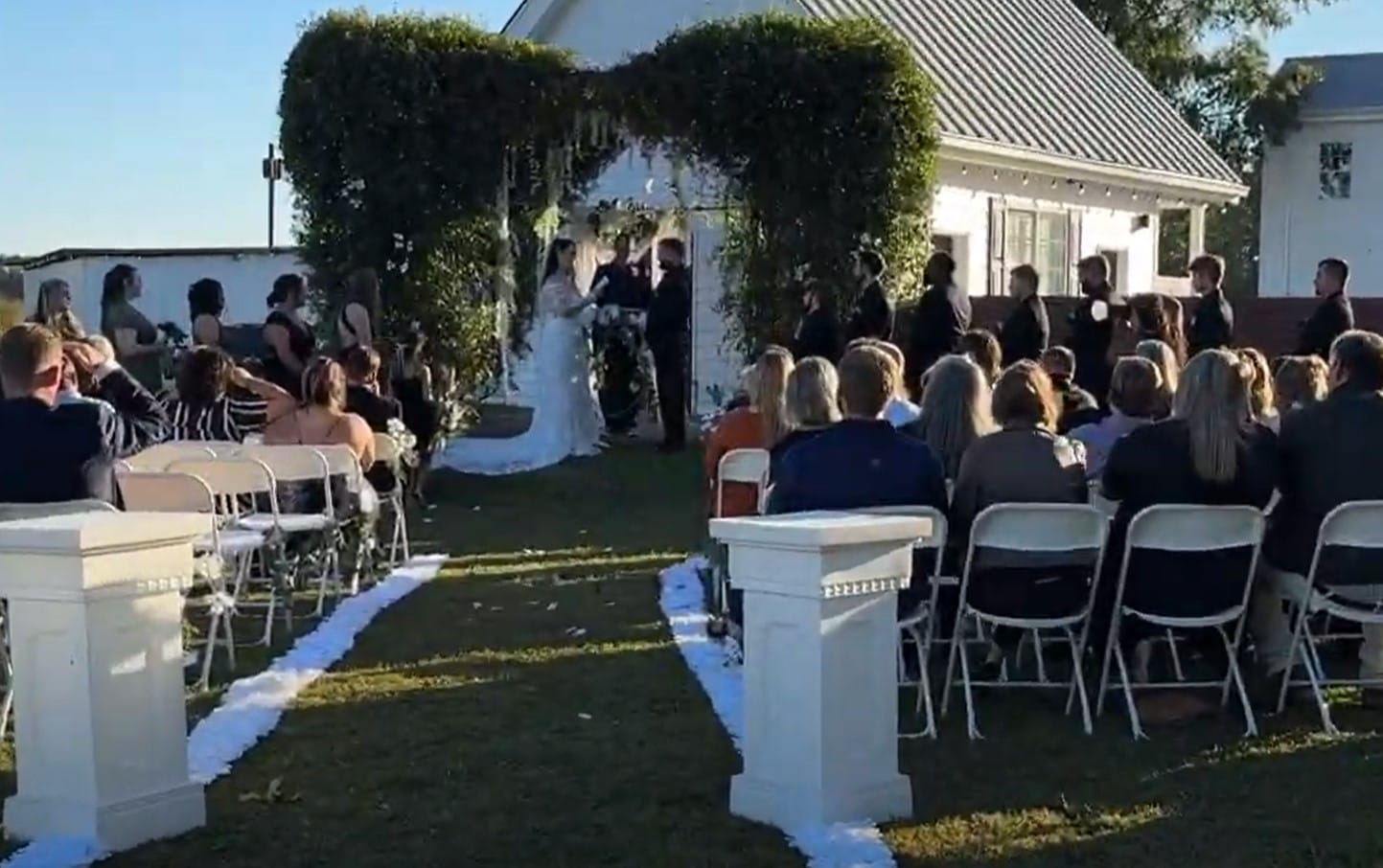 Outdoor wedding ceremony at Scarborough House with guests seated in white chairs along a white petal aisle leading to a lush greenery arch where the couple exchanges vows beside a historic white manor.