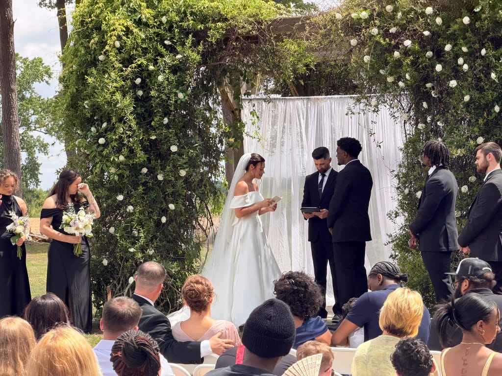 Bride in an off-shoulder gown reads her vows beneath a lush floral and greenery arch with white drapery at an outdoor wedding ceremony at Scarborough House.