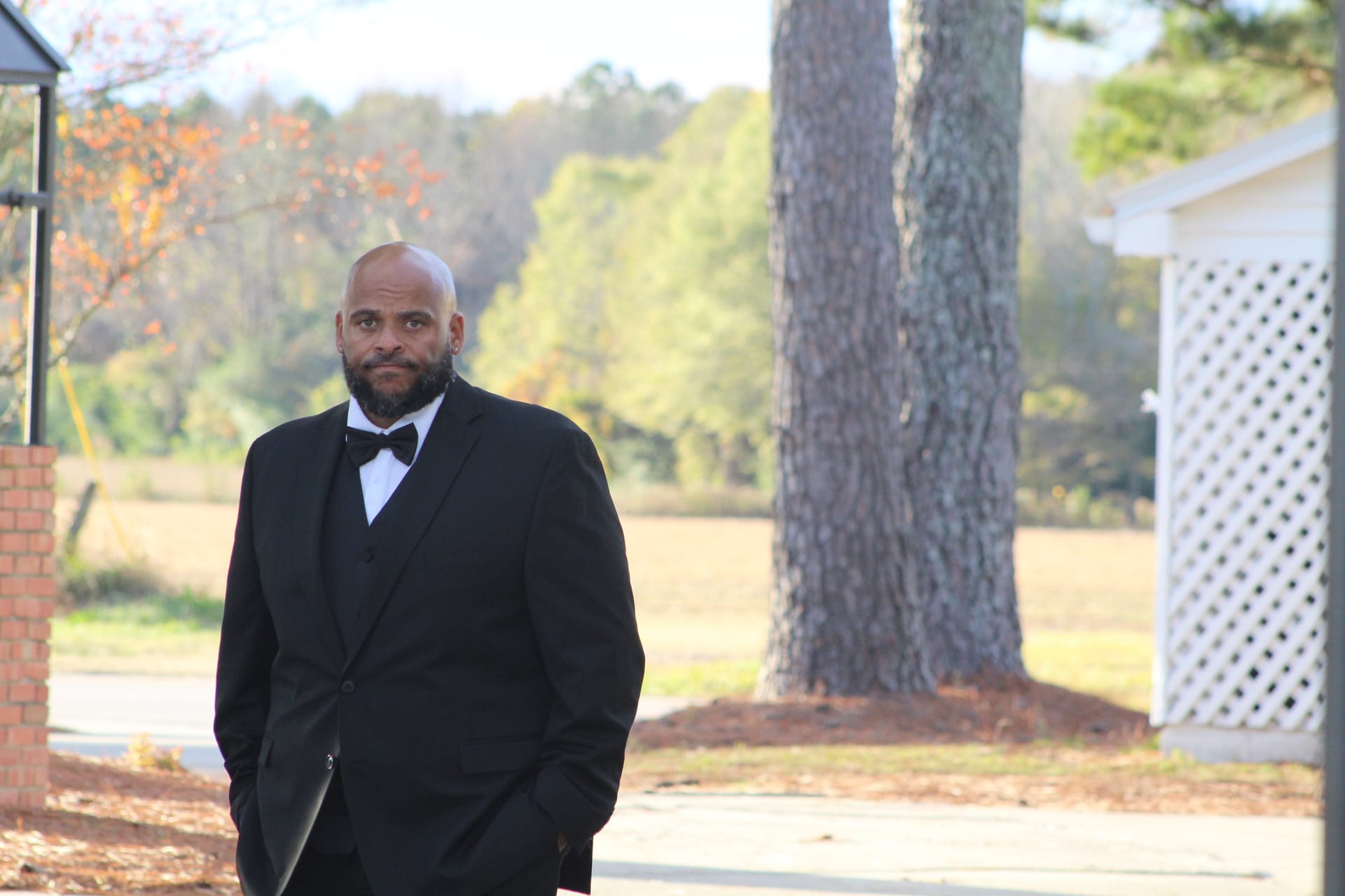 A bald groom with a beard dressed in a black tuxedo and bow tie stands outdoors near a large pine tree with open farmland fields visible in the background at Scarborough House estate.