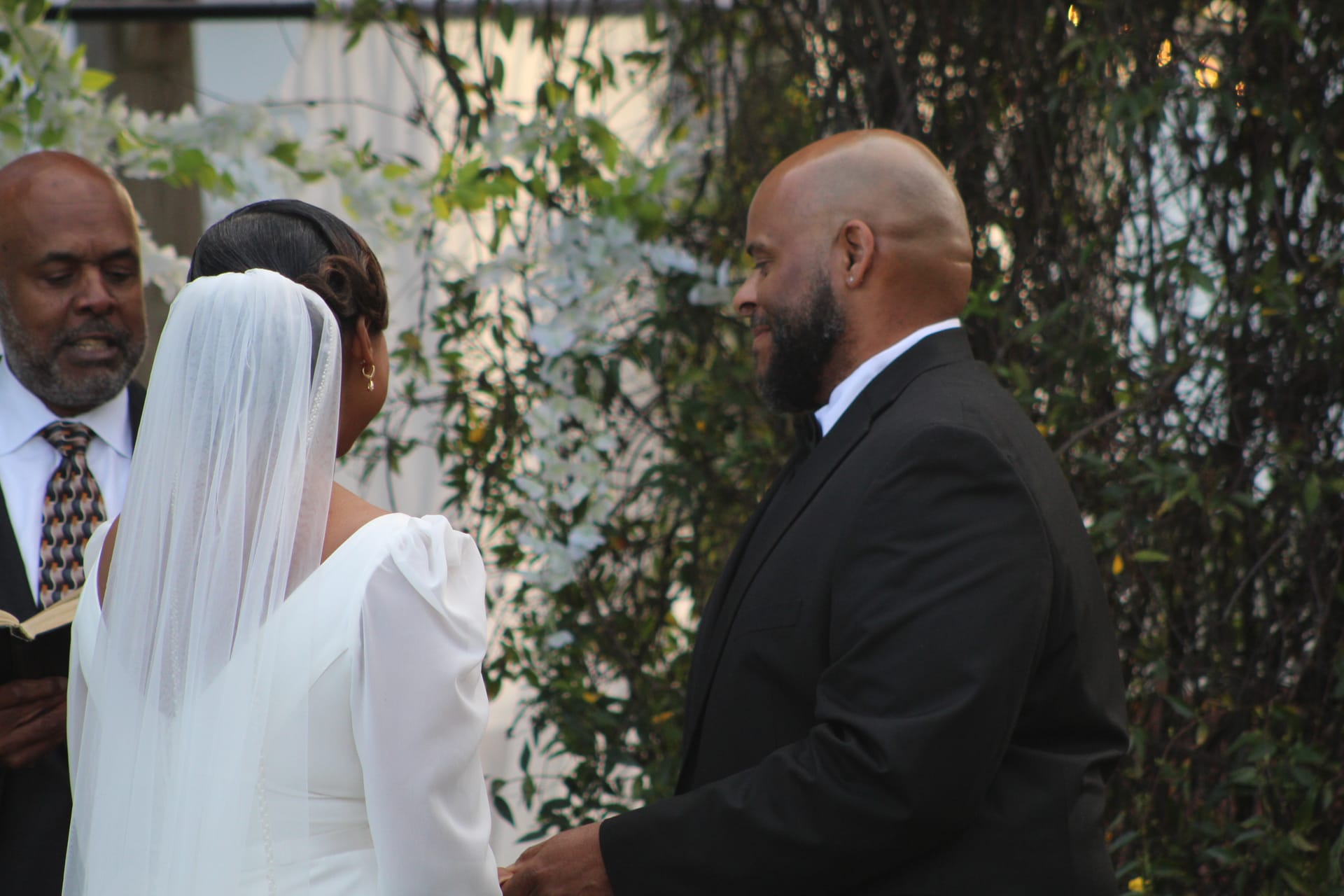 A groom in a black suit smiles tenderly at his bride in a white gown and veil during an outdoor wedding ceremony as the officiant reads from a book amid lush greenery.
