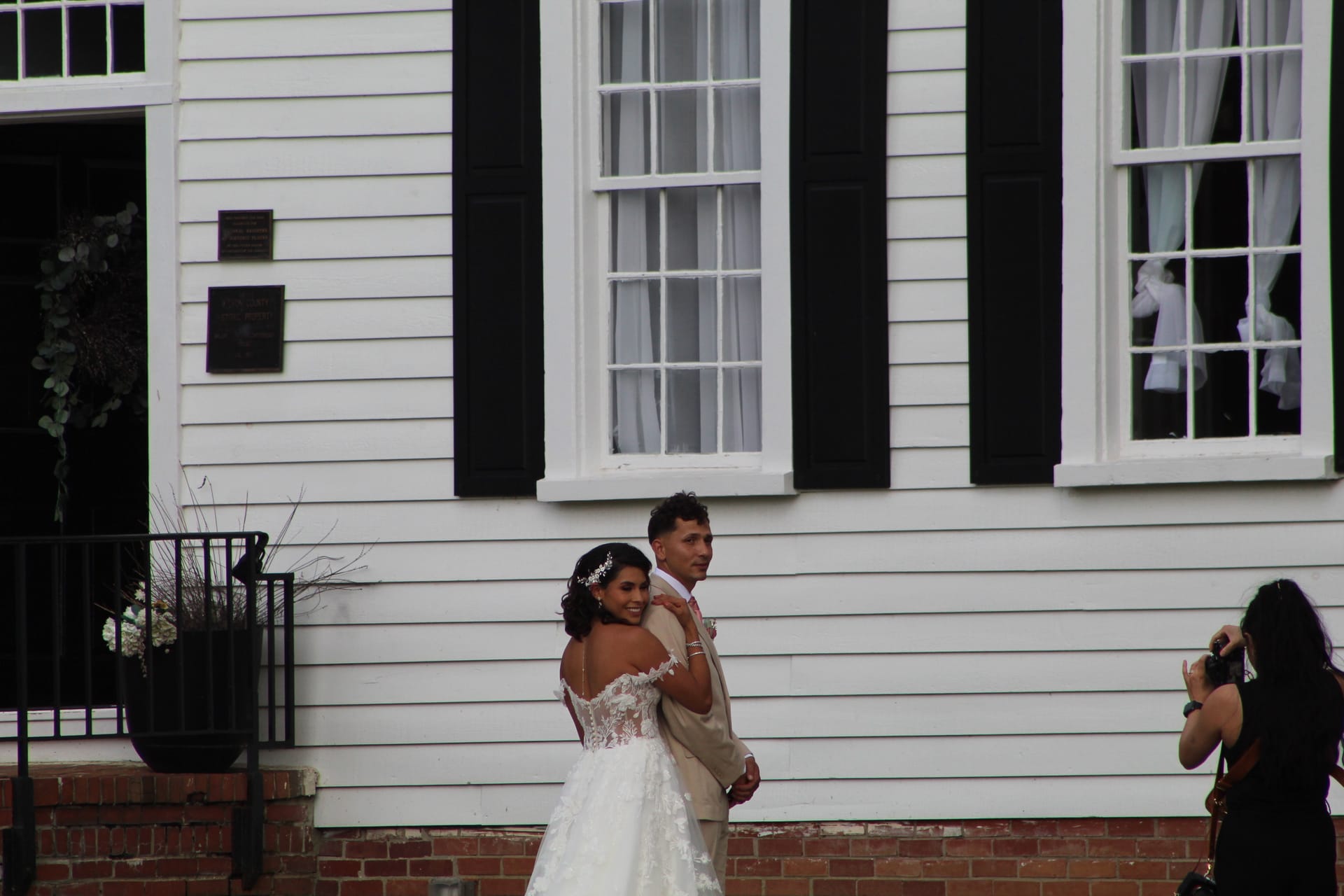 Bride in a lace off-the-shoulder gown and groom in a tan suit embrace and smile in front of the white clapboard facade of the 1821 Scarborough House manor while a photographer captures the moment.