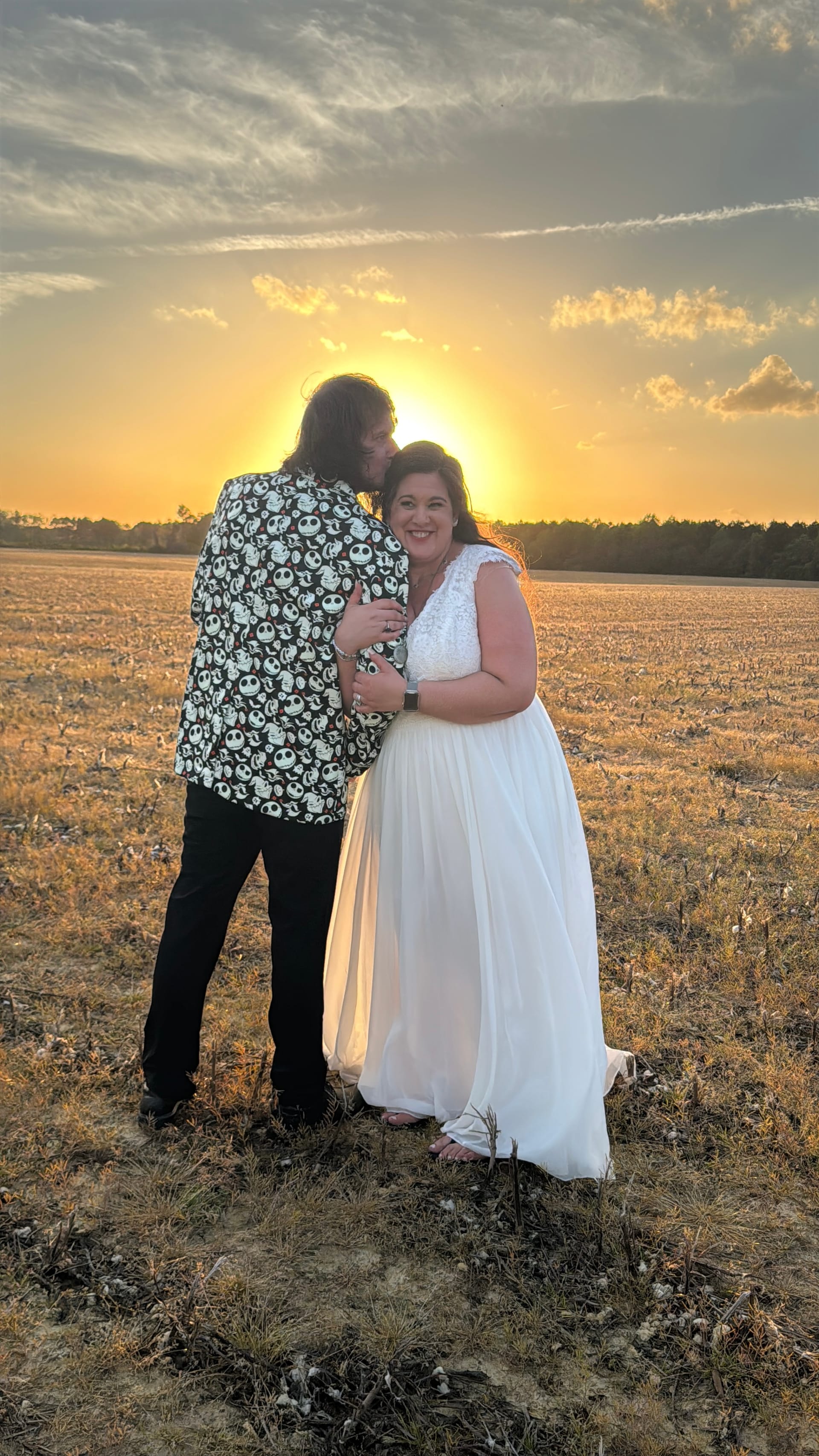 Bride in a flowing white gown smiles over her shoulder as her groom kisses her cheek during a golden sunset over the historic Scarborough House farmland fields.