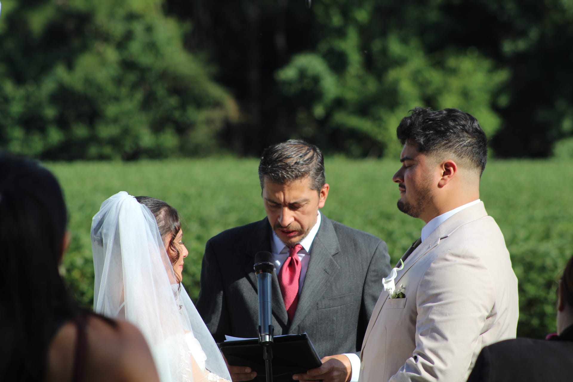 Bride in white veil and groom in a tan suit exchange vows before an officiant in a gray suit during an outdoor wedding ceremony surrounded by lush green fields and trees.