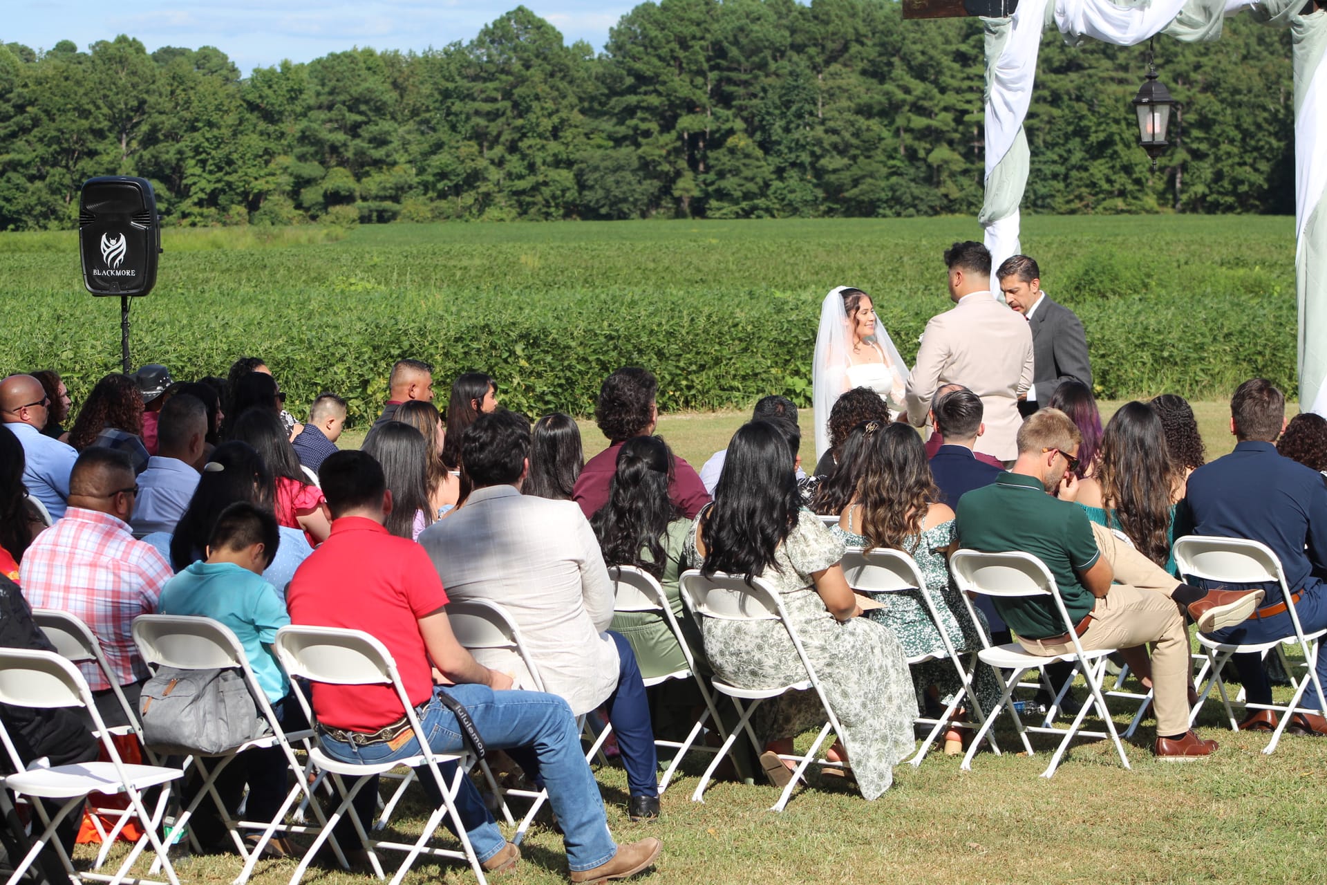 Wedding guests seated in white folding chairs on the ceremony lawn at Scarborough House watch as a bride in a white veil and groom exchange vows before an officiant, with lush green farmland fields and a wooded tree line stretching across the horizon behind them.