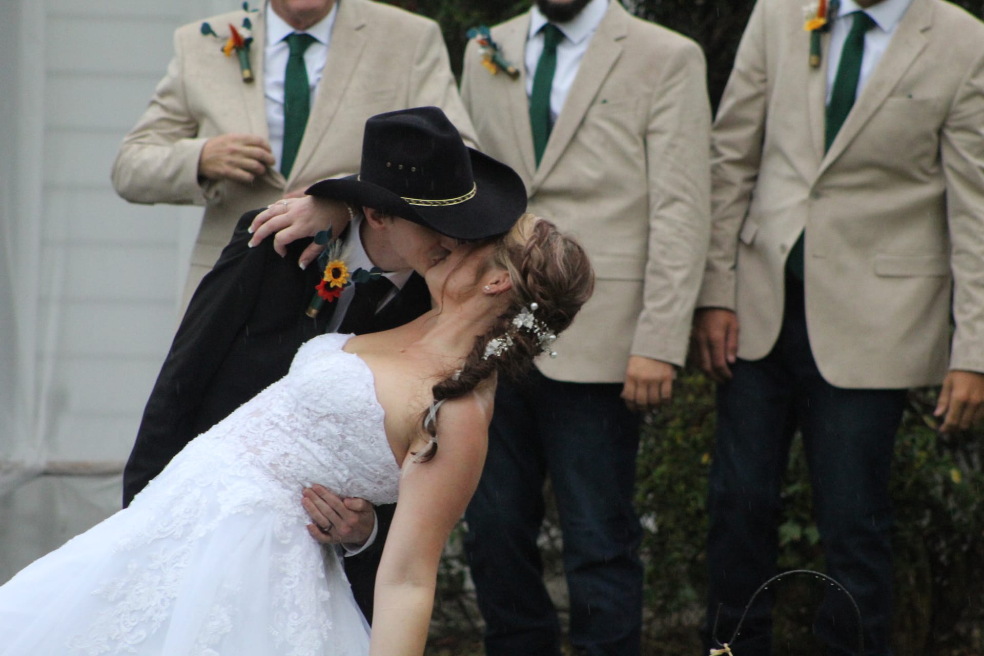 Groom in black cowboy hat dips bride in white lace gown for a kiss while groomsmen in tan blazers and green ties stand behind them at Scarborough House wedding venue.
