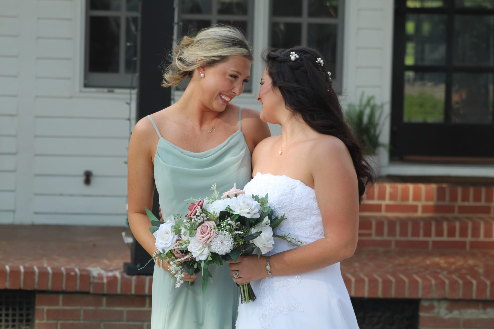 Bride in white strapless lace gown and bridesmaid in sage green dress share a joyful smile while holding floral bouquets outside the 1821 Scarborough House brick manor.