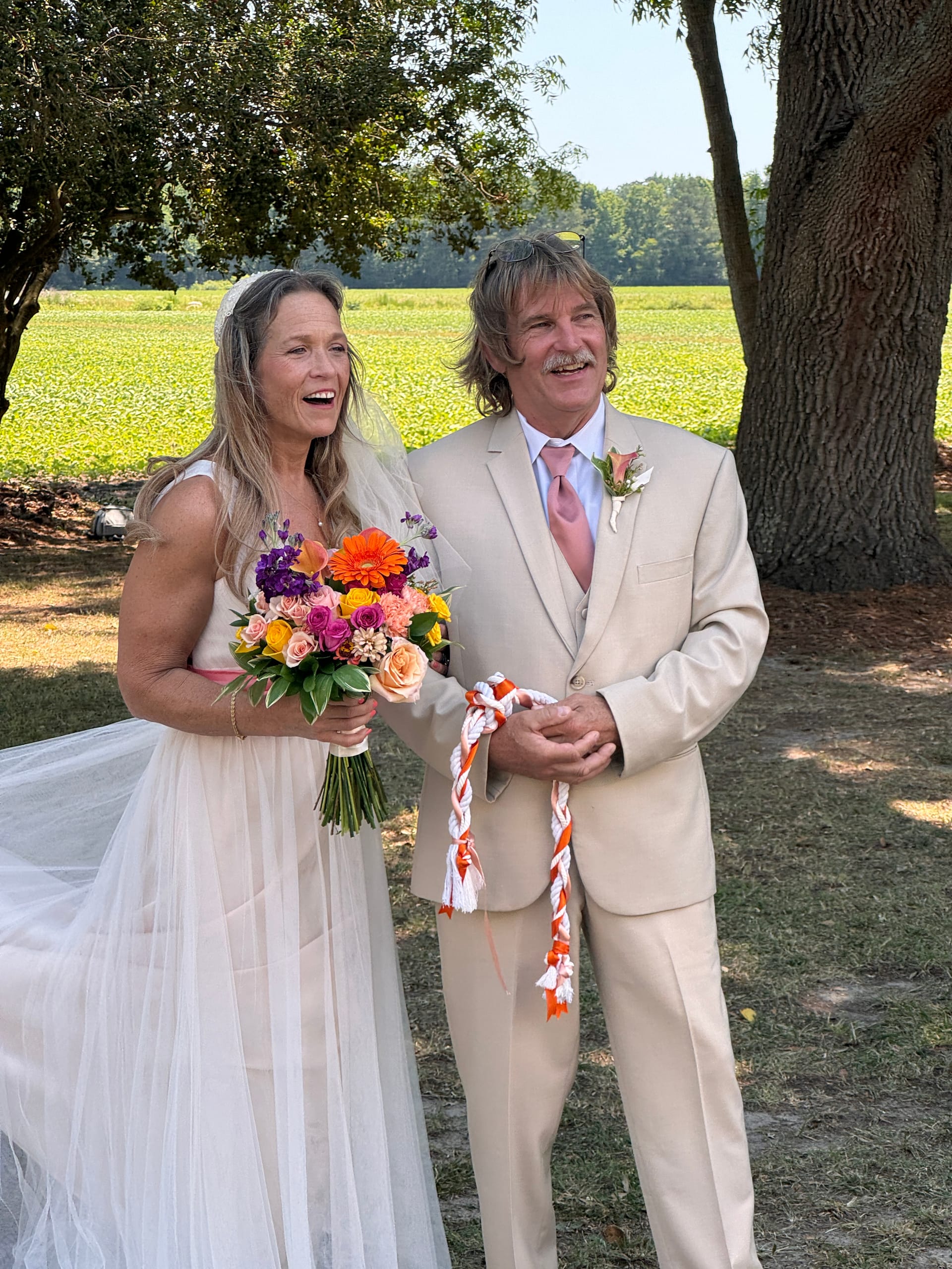 Laughing bride holding a colorful orange and pink bouquet stands beside her groom in a tan suit during a handfasting ceremony on the sun-drenched lawn at Scarborough House with green farmland fields behind them.