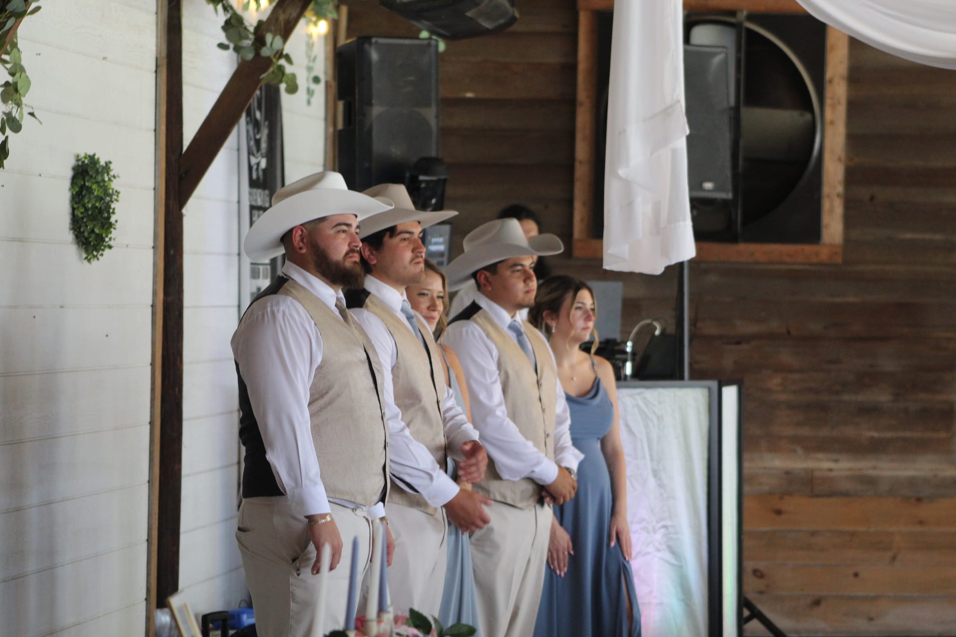 Groomsmen wearing tan vests and white cowboy hats stand attentively alongside bridesmaids in dusty blue gowns during an indoor barn-style wedding ceremony.