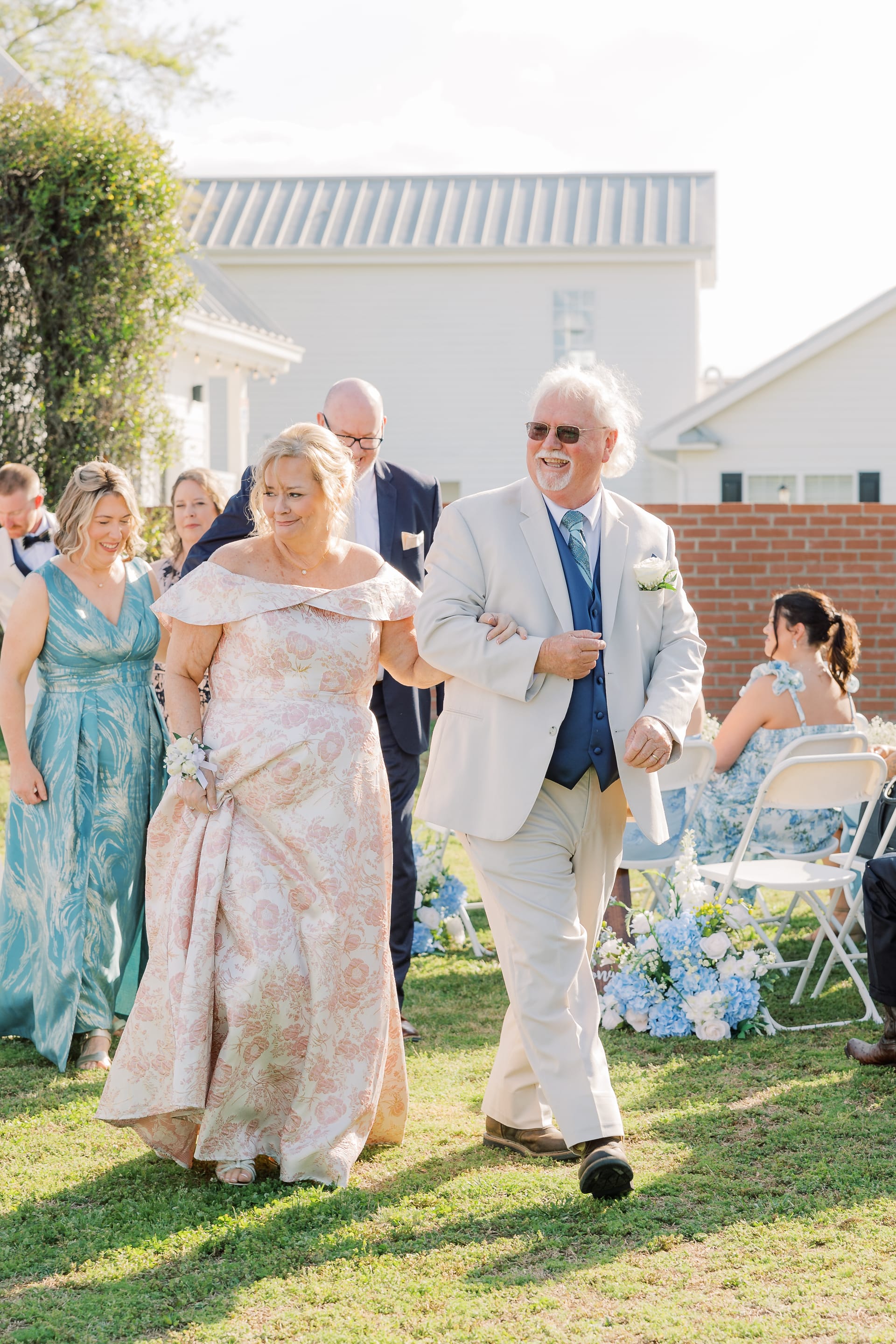 A smiling couple walks arm in arm down the outdoor ceremony aisle at Scarborough House, surrounded by blue hydrangea floral arrangements and wedding guests seated on white folding chairs.