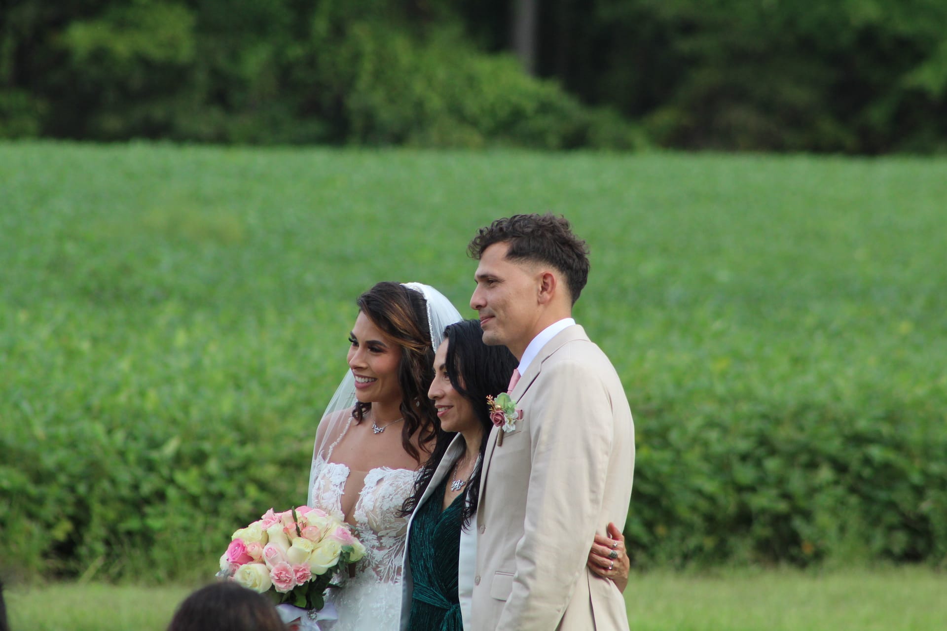 Smiling bride in lace gown holding pink and white bouquet stands with groom in tan suit beside a guest on the ceremony lawn with green farmland fields in the background at Scarborough House.