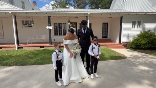 Bride in an off-shoulder white gown holding a bouquet stands with her groom in a dark suit and two young boys in suspenders and bow ties in front of the historic 1821 Scarborough House manor.