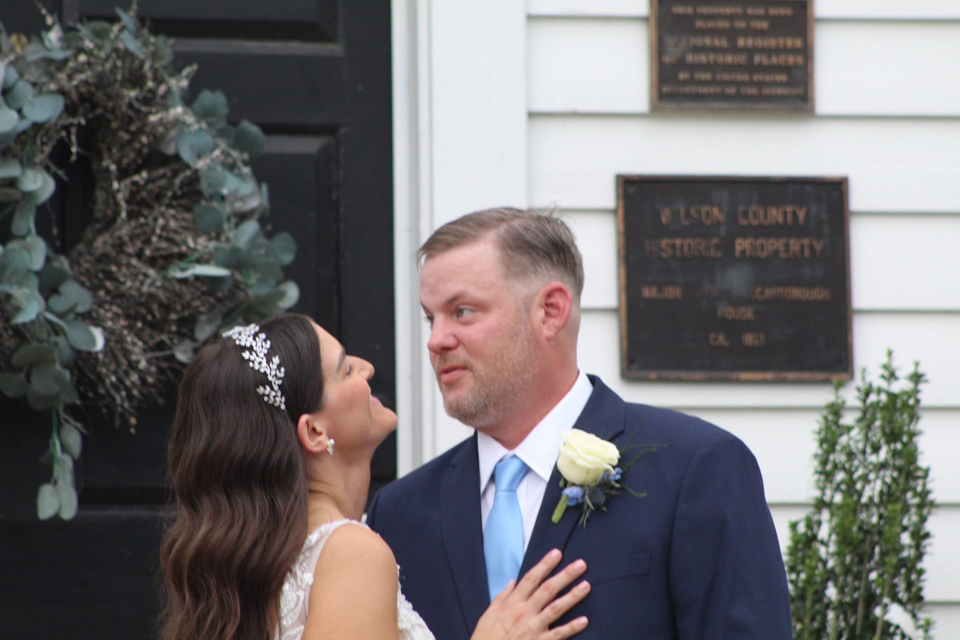 Bride in lace gown and floral crown gazes up at groom in navy suit with light blue tie and boutonniere outside the 1821 Scarborough House historic manor entrance.