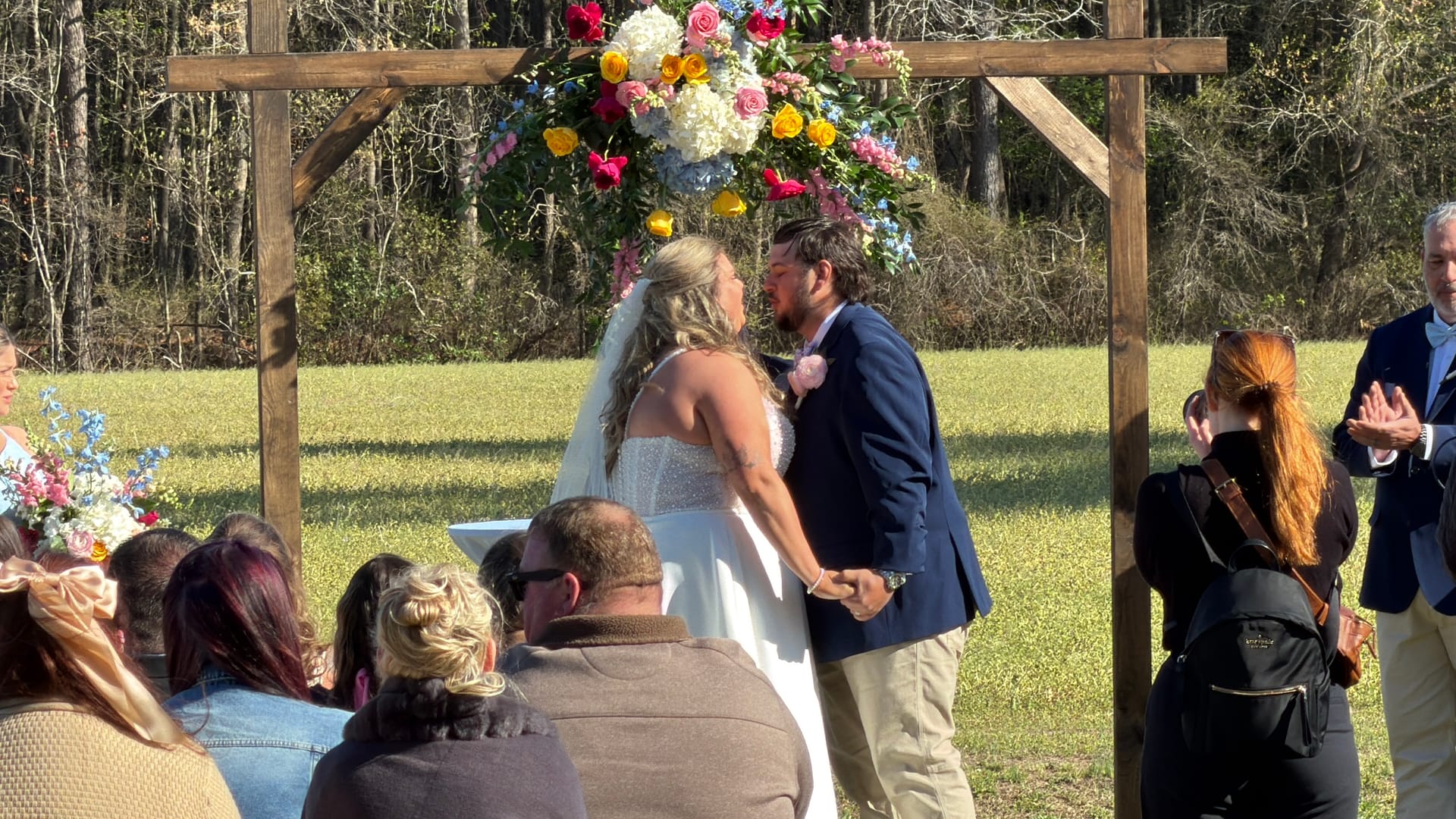 Bride and groom share their first kiss beneath a rustic wooden arch adorned with colorful pink yellow and white floral arrangements on the ceremony lawn surrounded by seated wedding guests.