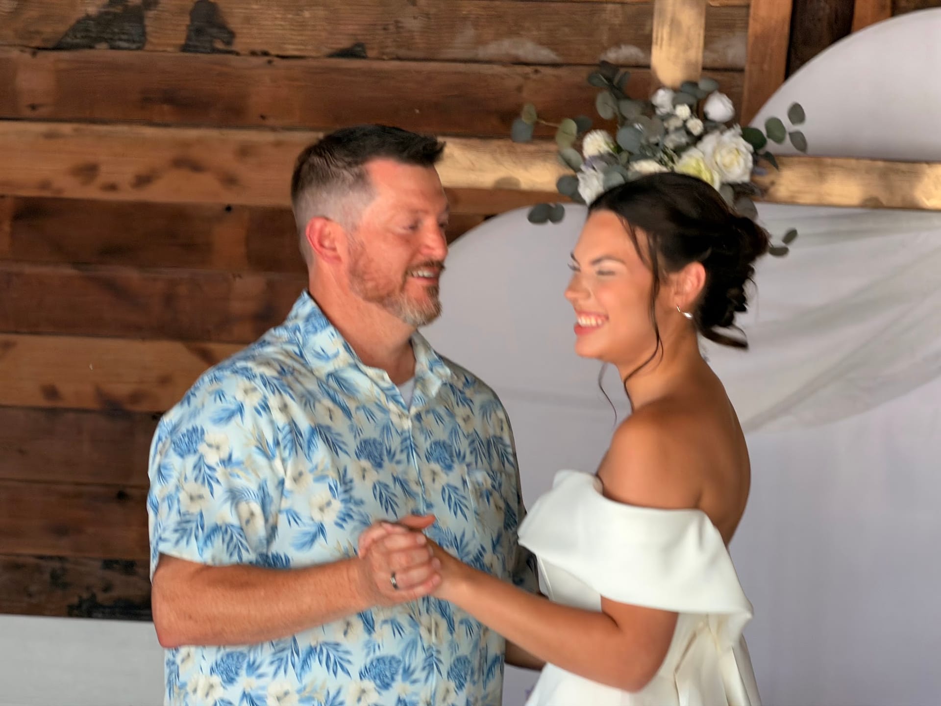 A smiling bride in a white off-shoulder gown and floral crown shares a first dance with the groom in a blue floral shirt against a rustic wood-plank wall at Scarborough House.