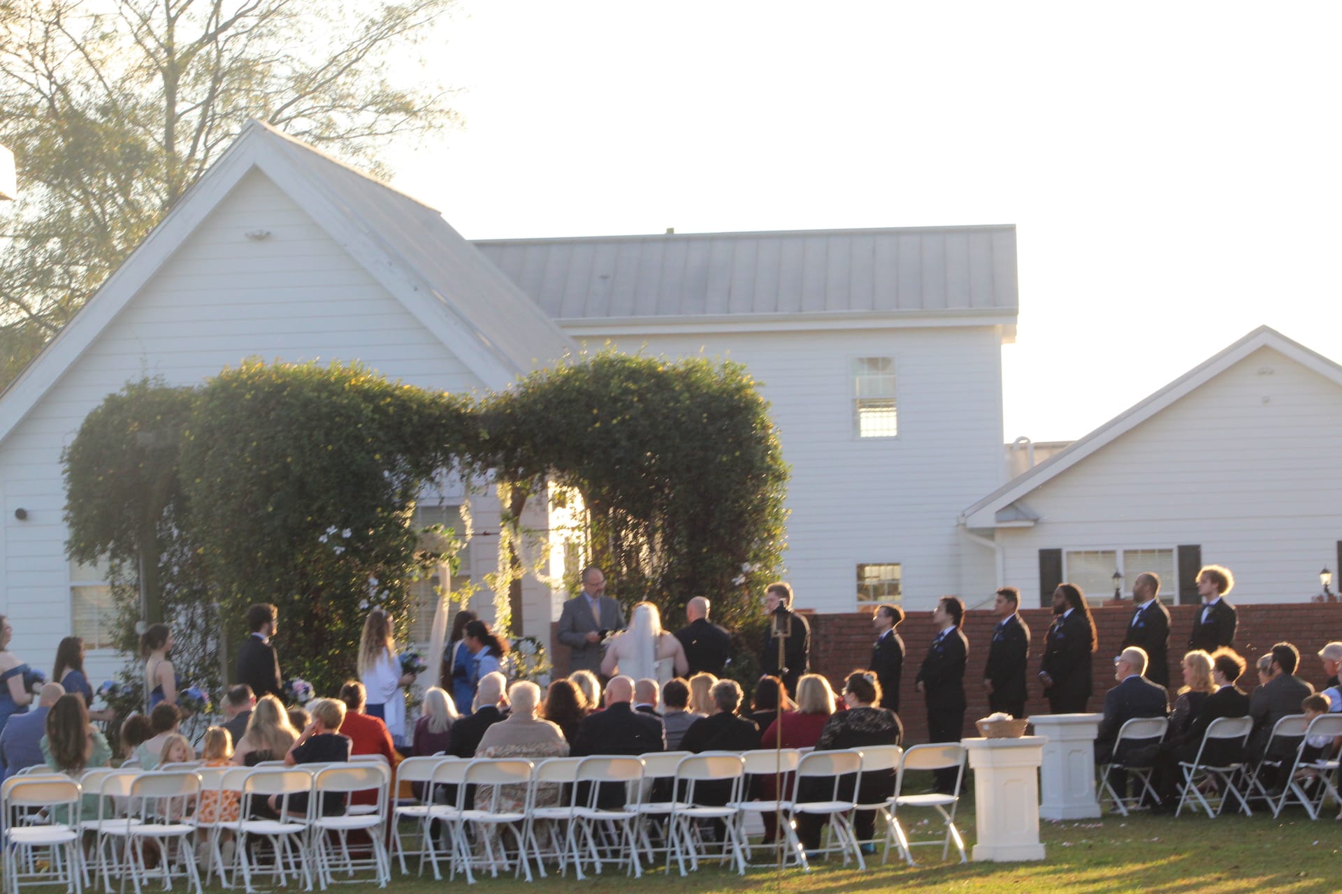 Outdoor wedding ceremony on the lawn of a historic white manor house with guests seated in white folding chairs facing the couple under a lush ivy-covered archway at golden hour.