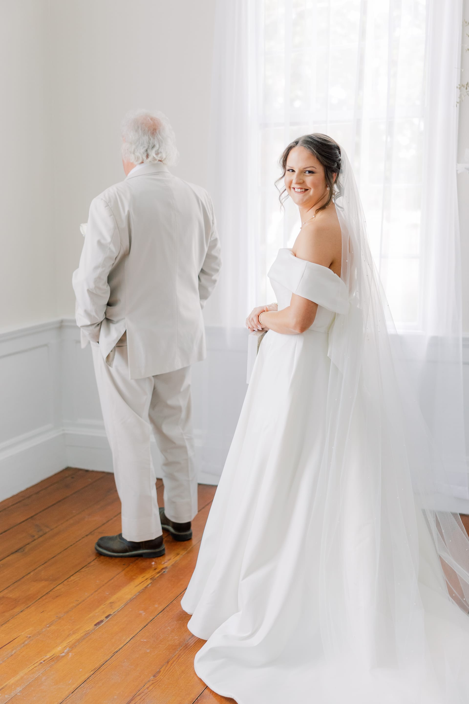 A smiling bride in an off-shoulder white gown and veil glances back at the camera while an older gentleman in a light suit stands with his back turned during a first look moment inside a bright historic room at Scarborough House.