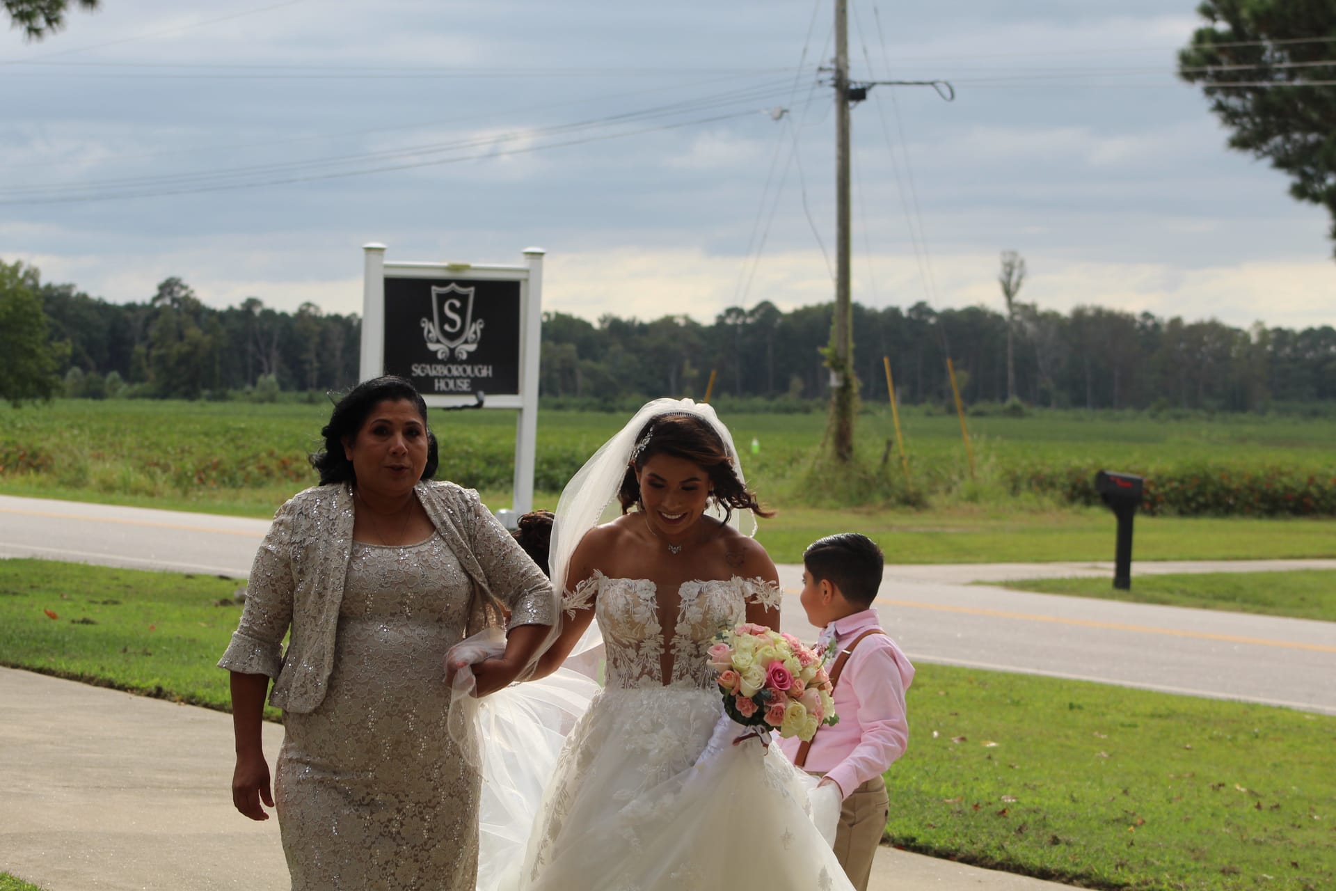 A smiling bride in a lace off-shoulder gown carries a pink and white bouquet while walking with a woman in a silver dress and a young boy in pink toward Scarborough House on her wedding day.