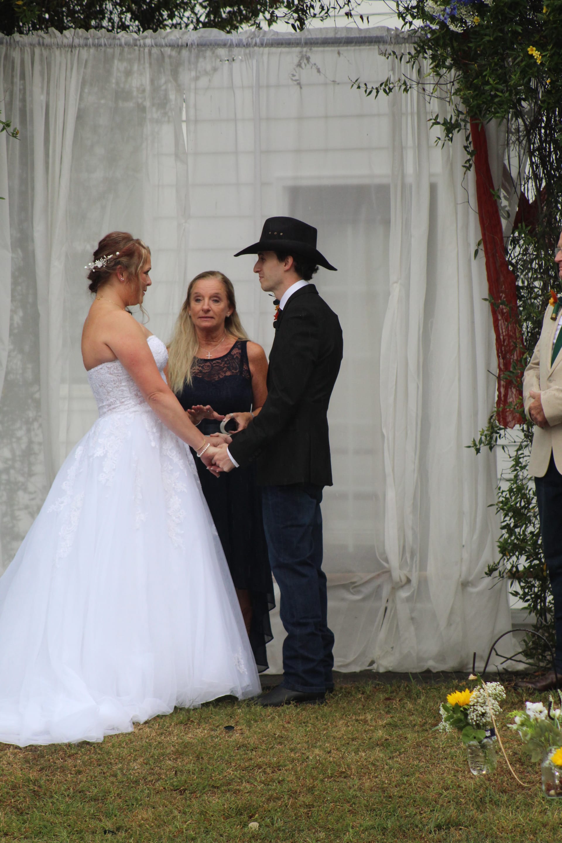 Bride in a white ball gown and groom in a black cowboy hat hold hands during their outdoor wedding ceremony before a sheer white draped backdrop at Scarborough House.